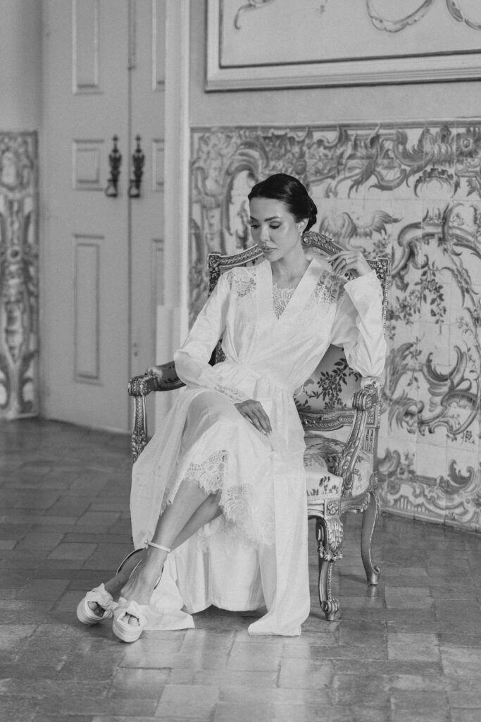 The bride in a white robe adjusts her shoe, seated by an azulejo tile panel at Palácio do Correio-Mor