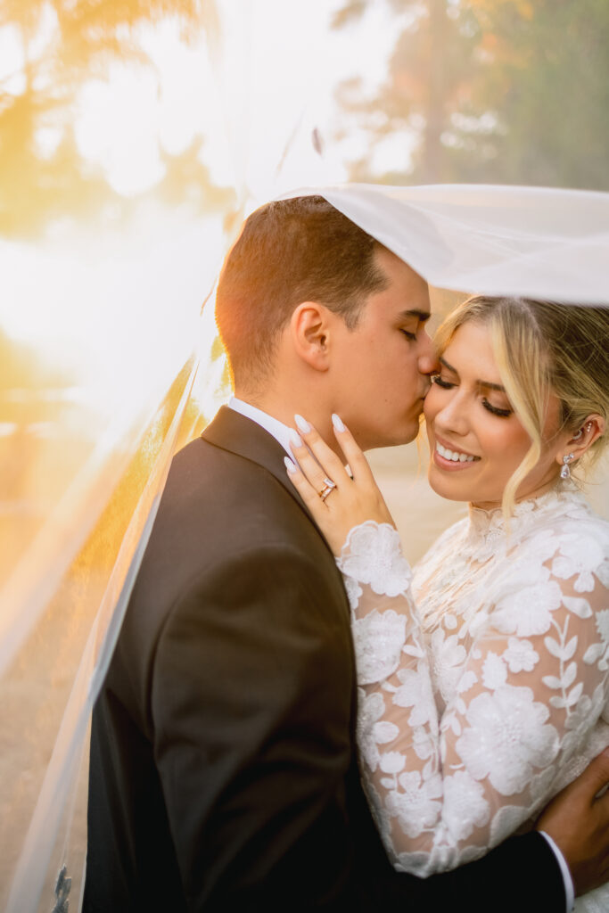 The newlyweds beneath the veil in golden late-afternoon light, the groom kissing the bride's forehead as she laughs