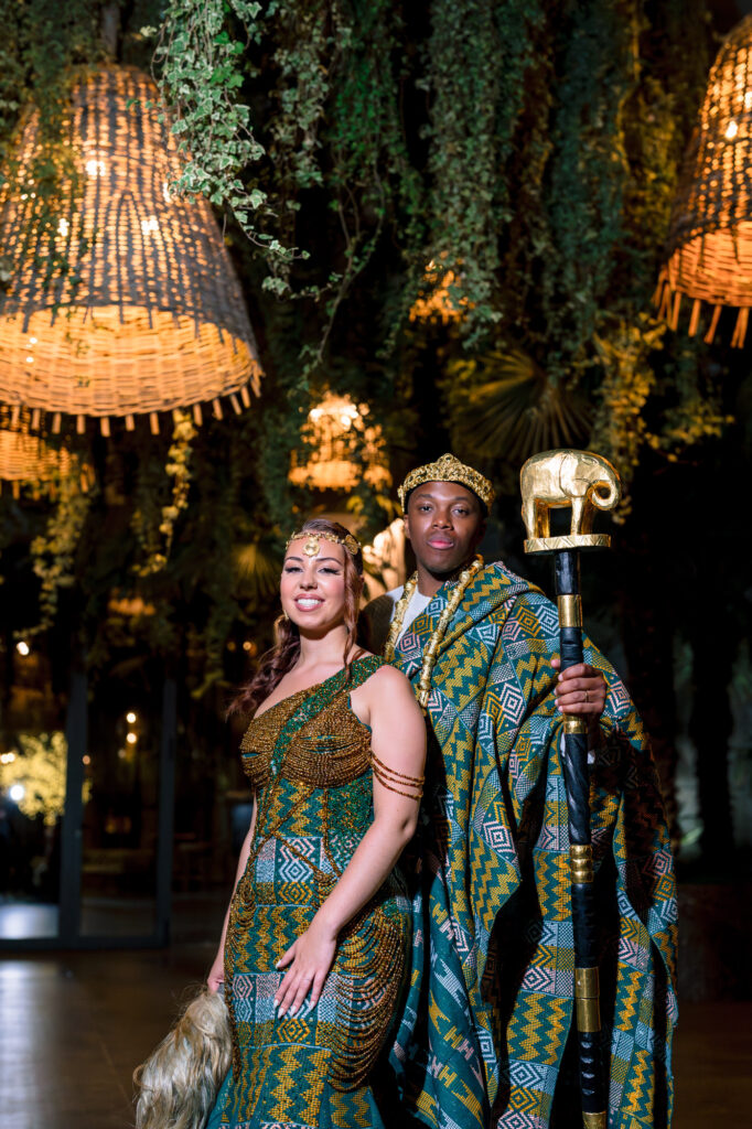 The newlyweds in traditional kente-patterned African attire beneath woven pendant lamps, the groom holding a gold elephant-topped ceremonial staff