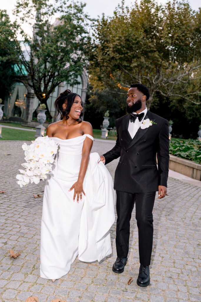 The bride and groom laugh together as they walk along a stone path at Solar da Levada, her bouquet of white orchids in hand