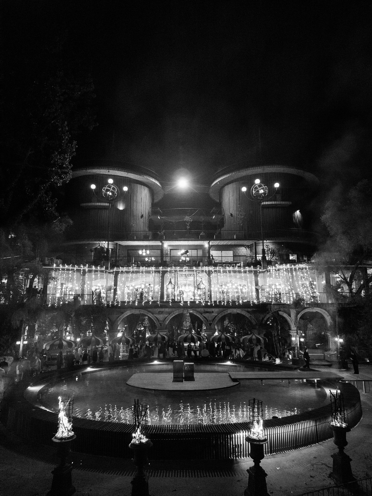 Night scene of a lit amusement park with a central fountain, string lights, and a two-tower building behind a carousel.