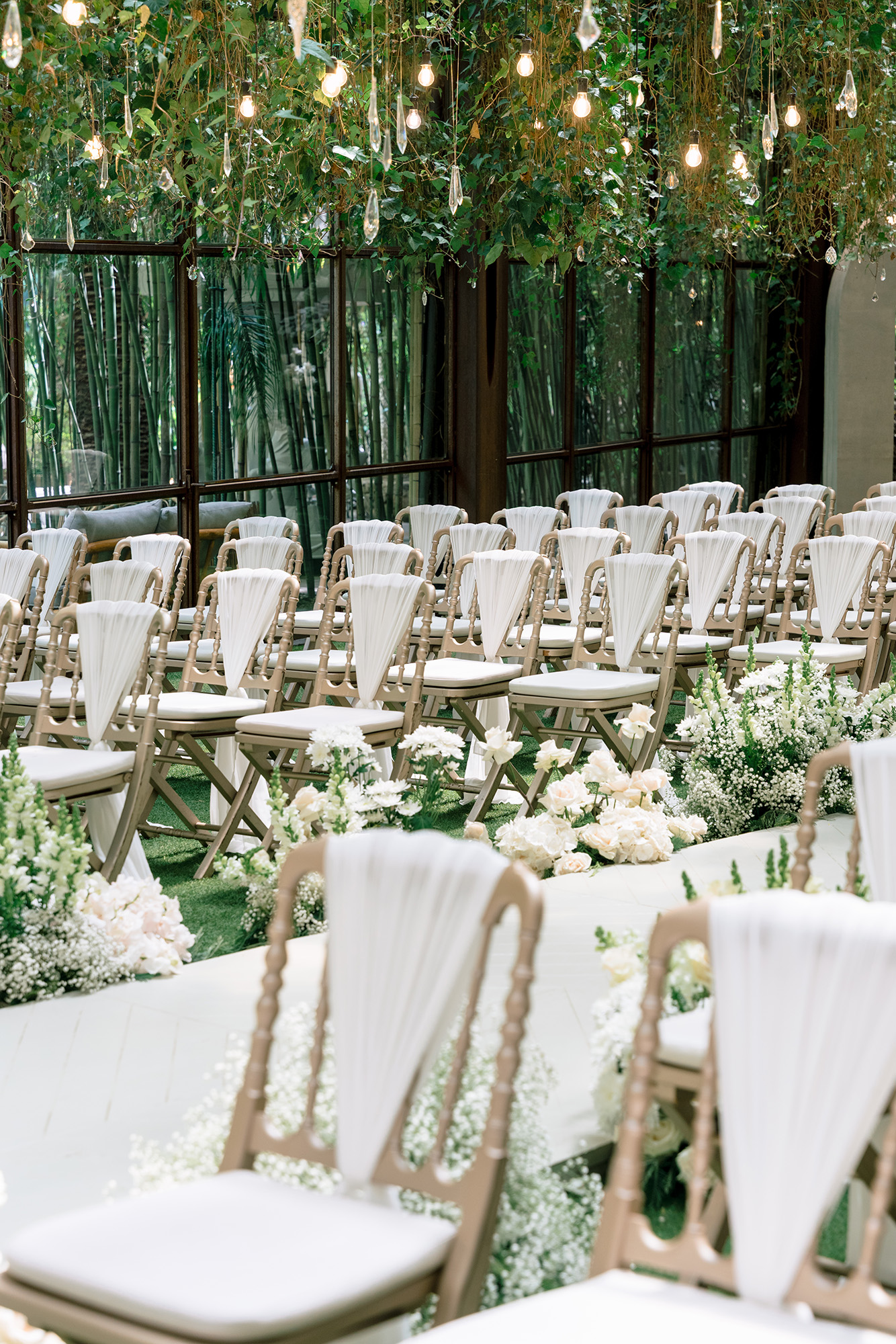 Rows of cream chairs with white fabric backs line an aisle, under lush greenery and warm hanging lights for a wedding ceremony.