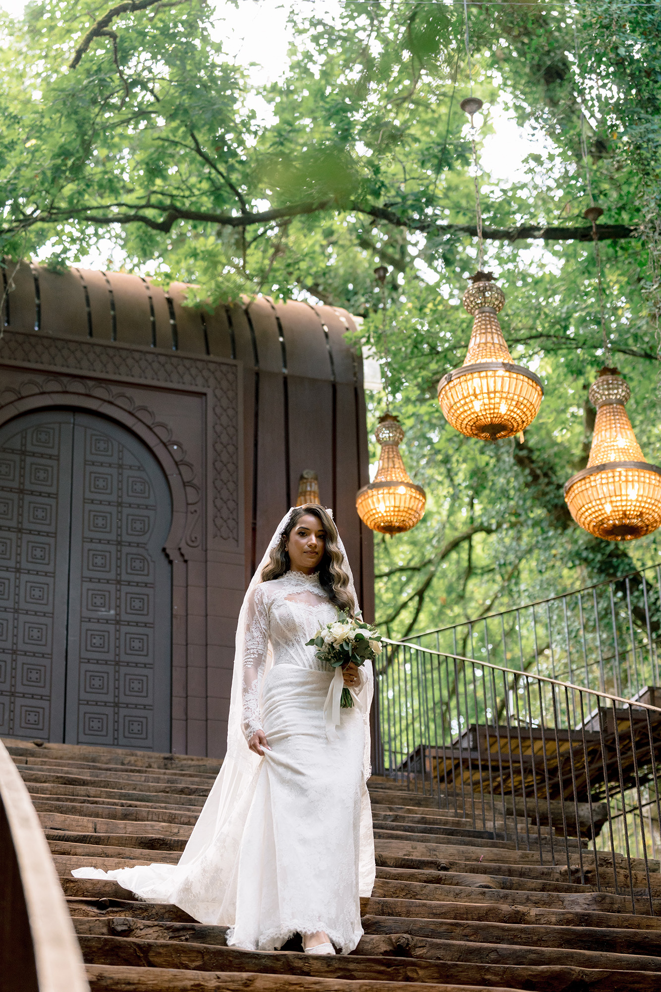 Bride in a lace white wedding gown holding a bouquet on wooden steps outdoors under hanging lanterns and green trees.