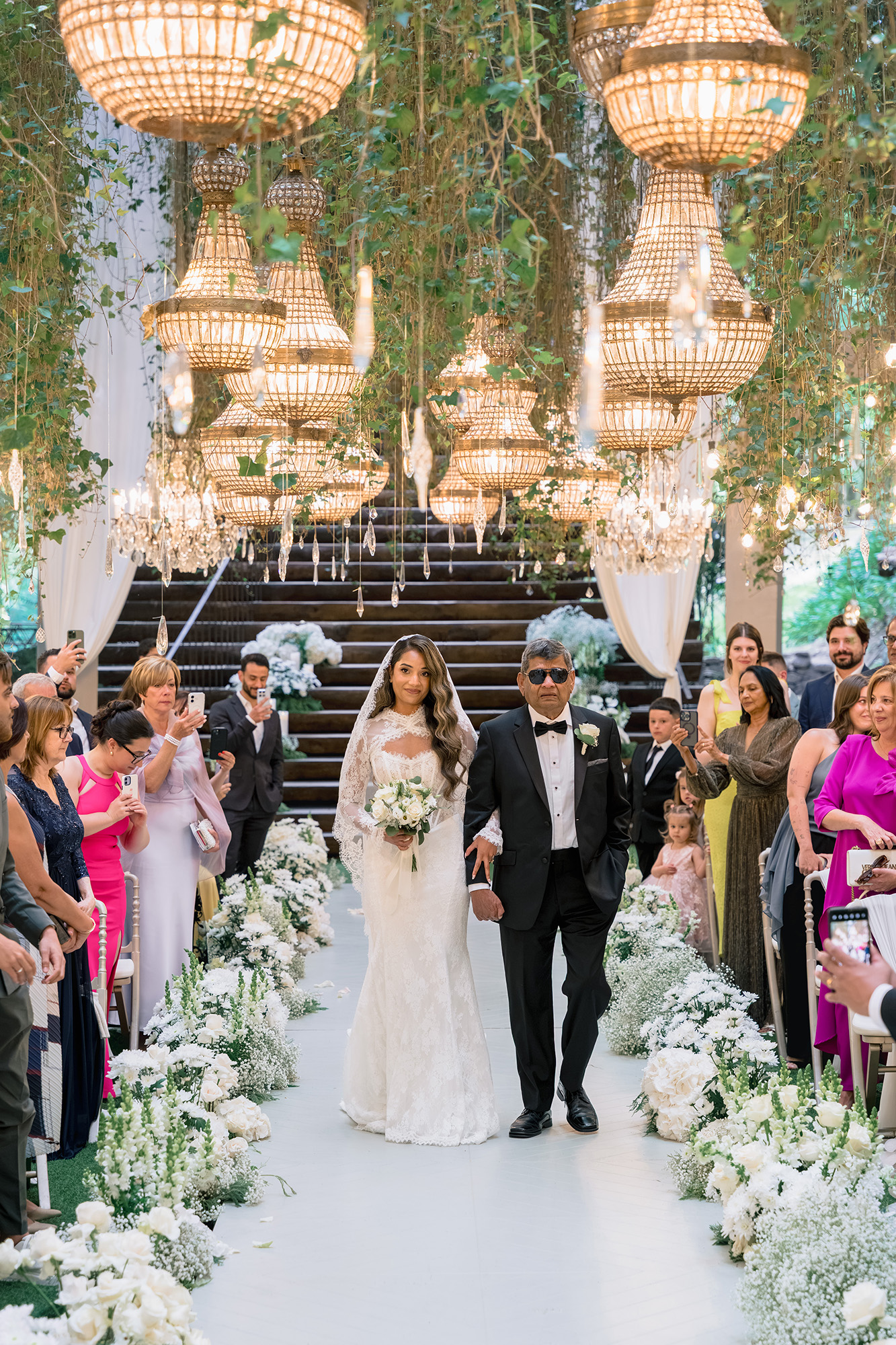 Bride in a white lace gown walks arm-in-arm with a man in a tuxedo down a white floral aisle beneath hanging chandeliers, as guests look on and take photos.