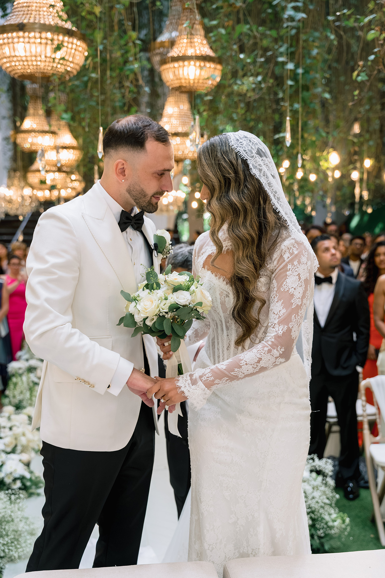 Bride and groom exchange rings during their wedding ceremony, with a bouquet of white flowers in the foreground and hanging chandeliers in the background.