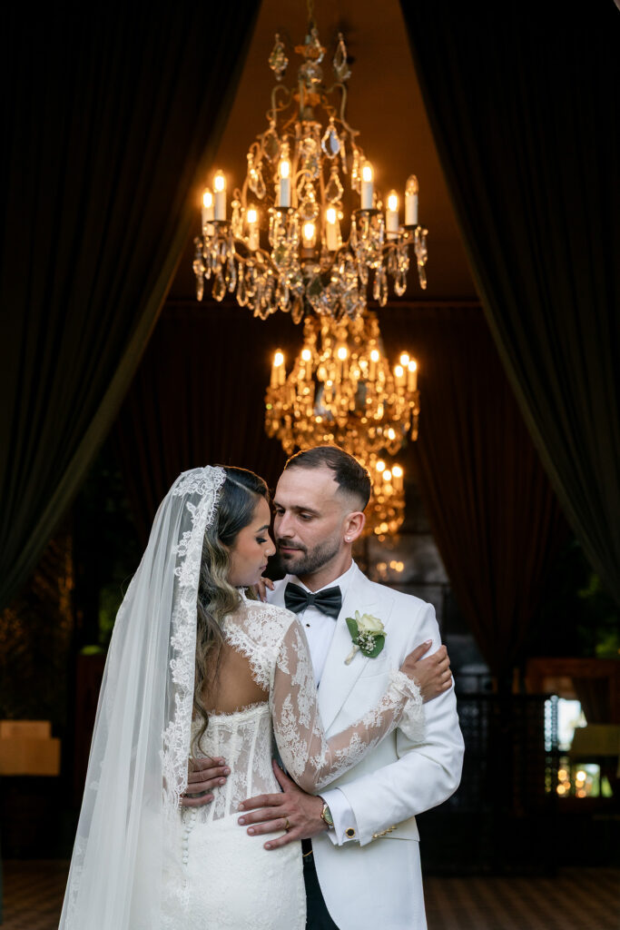 Couple in wedding attire embracing under a grand chandelier with warm candlelight glow behind them.