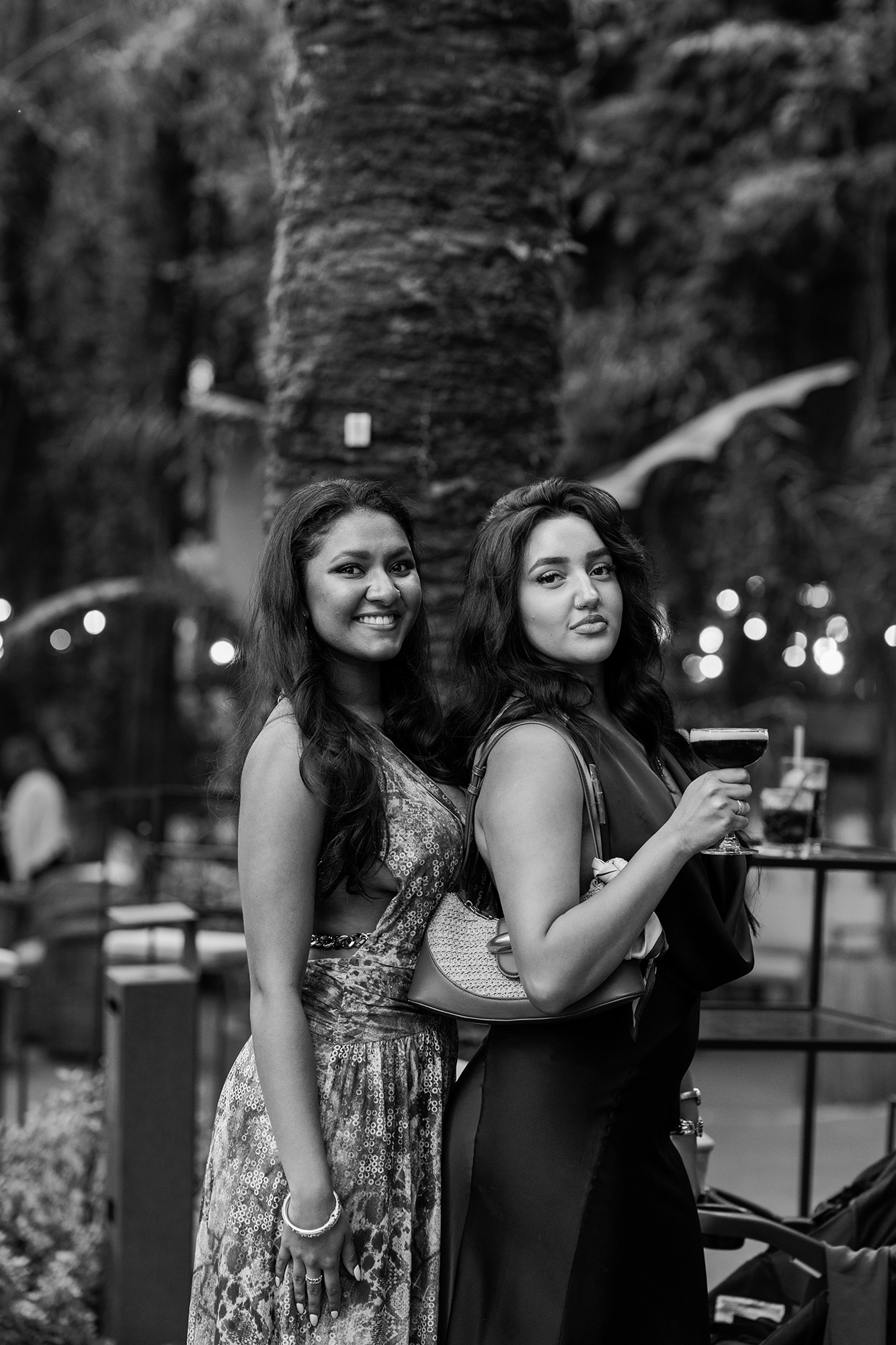 Two women posing outdoors at night, one smiling at the camera and the other holding a wine glass.