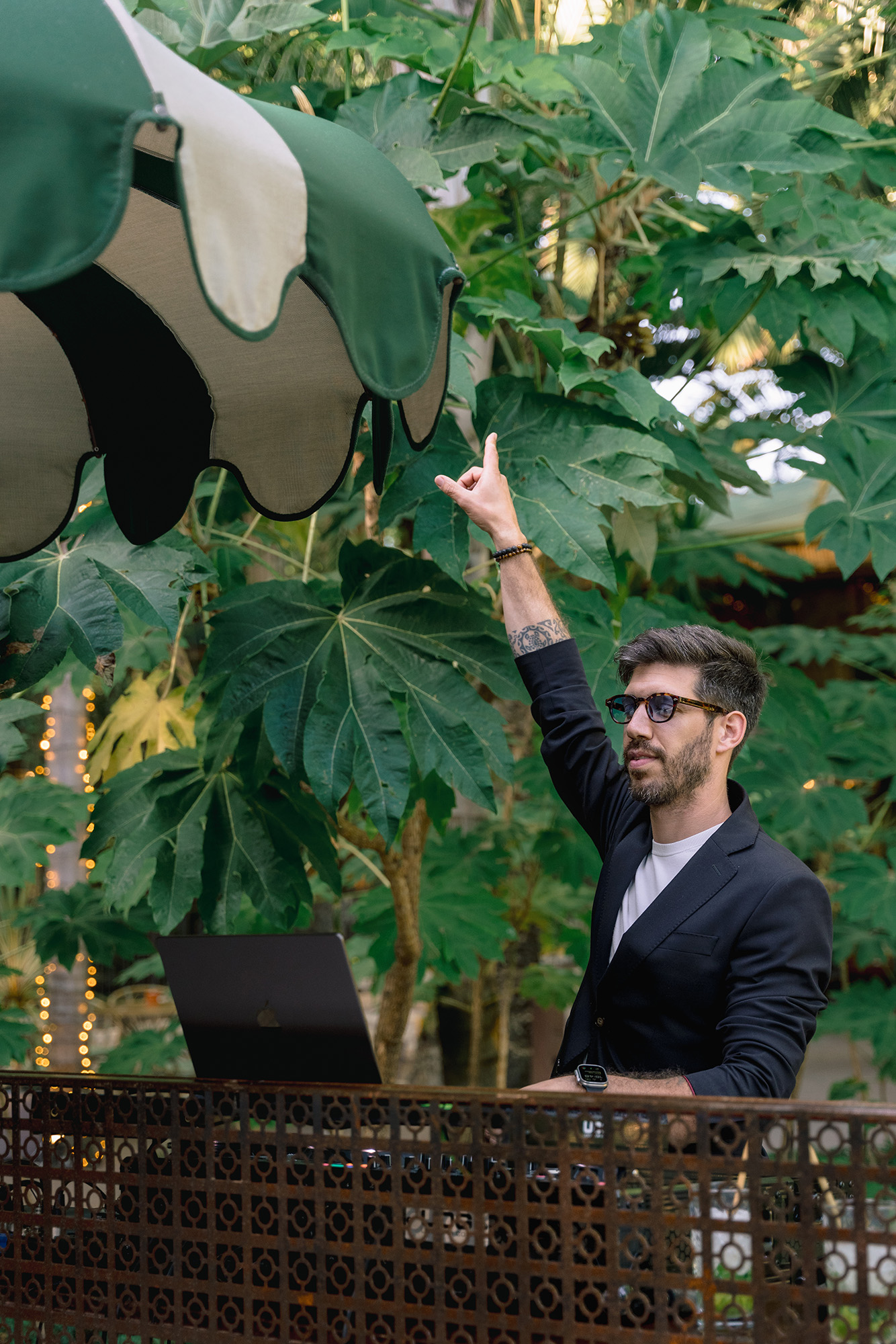 Man in a black blazer with sunglasses raises his hand toward a large green outdoor canopy, laptop on a table nearby.