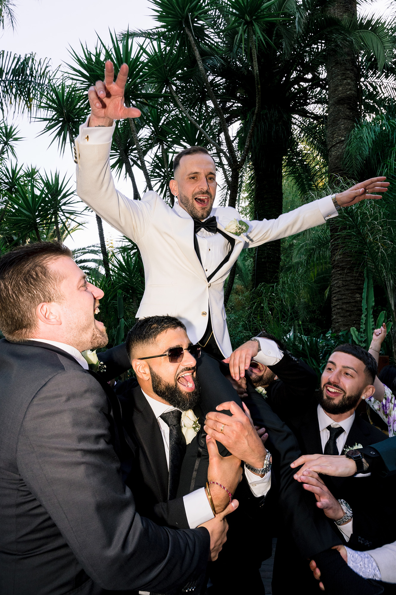 Groom in a white suit being carried on friends' shoulders at a wedding, smiling, with tropical trees in the background.