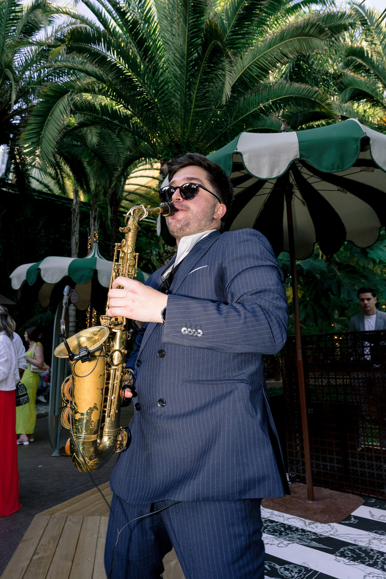 Jazz saxophonist in a navy pinstripe suit wearing sunglasses, playing outdoors at a garden party with palm trees nearby.