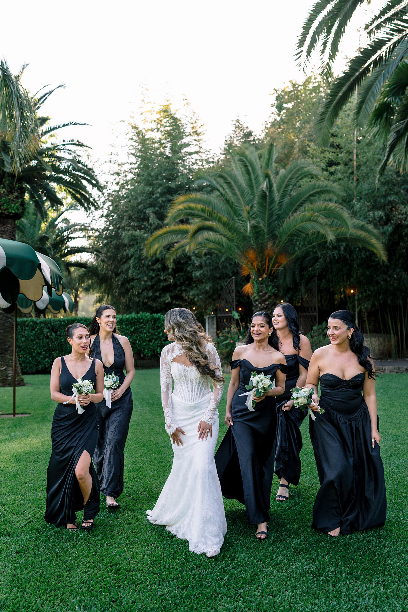 Bride in a white lace gown walks with bridesmaids in black dresses outdoors on a green lawn. nearby palm trees frame the scene.