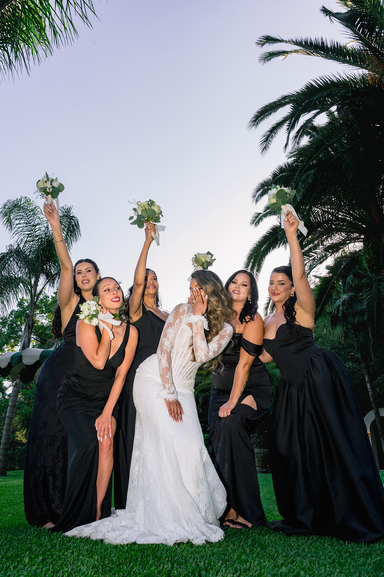 Bride in a white lace wedding gown surrounded by bridesmaids in black dresses, all raising small bouquets at sunset in a lush garden with palm trees.