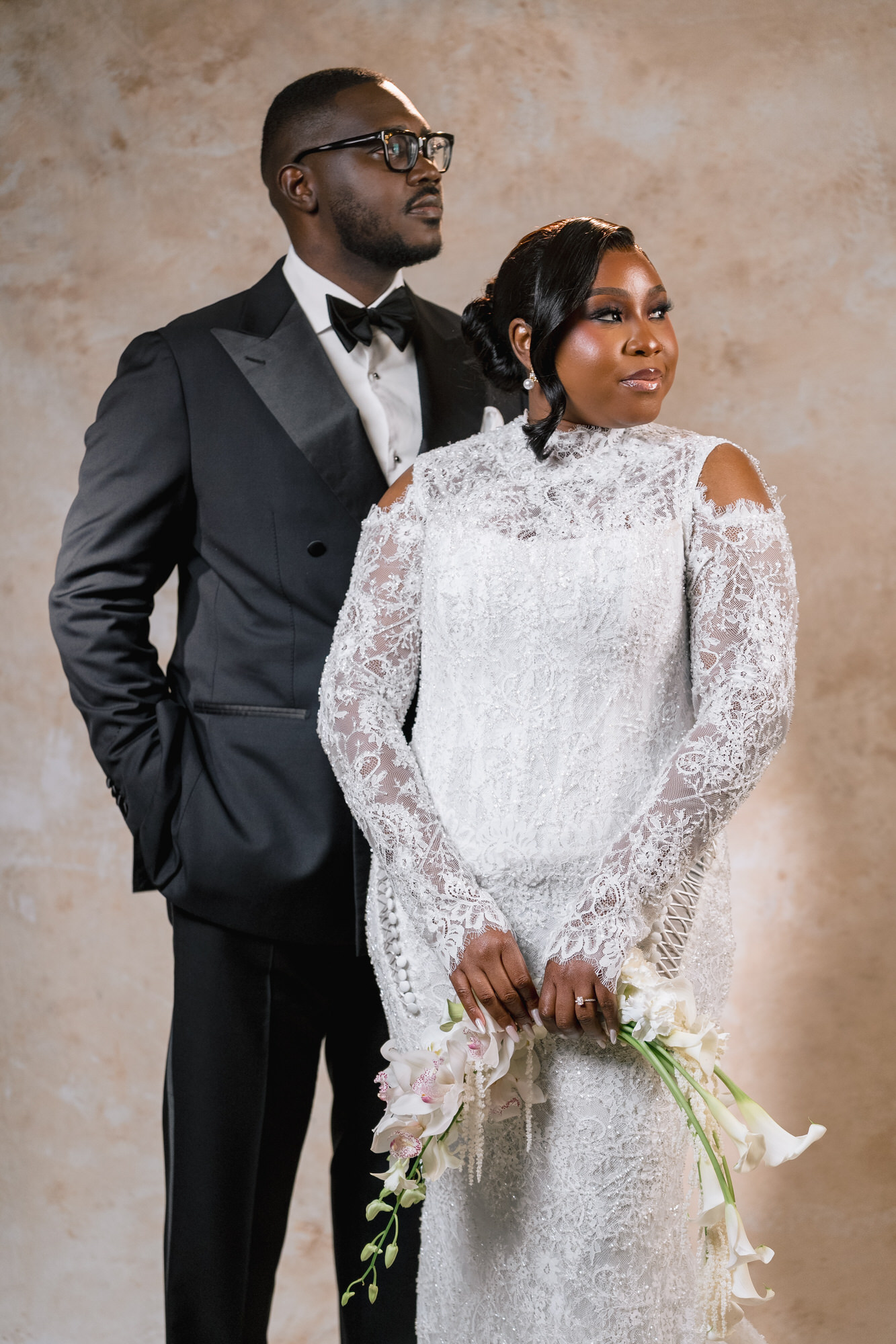 Bride in a white lace wedding gown holds a bouquet, with a groom in a black tuxedo beside her against a beige backdrop.