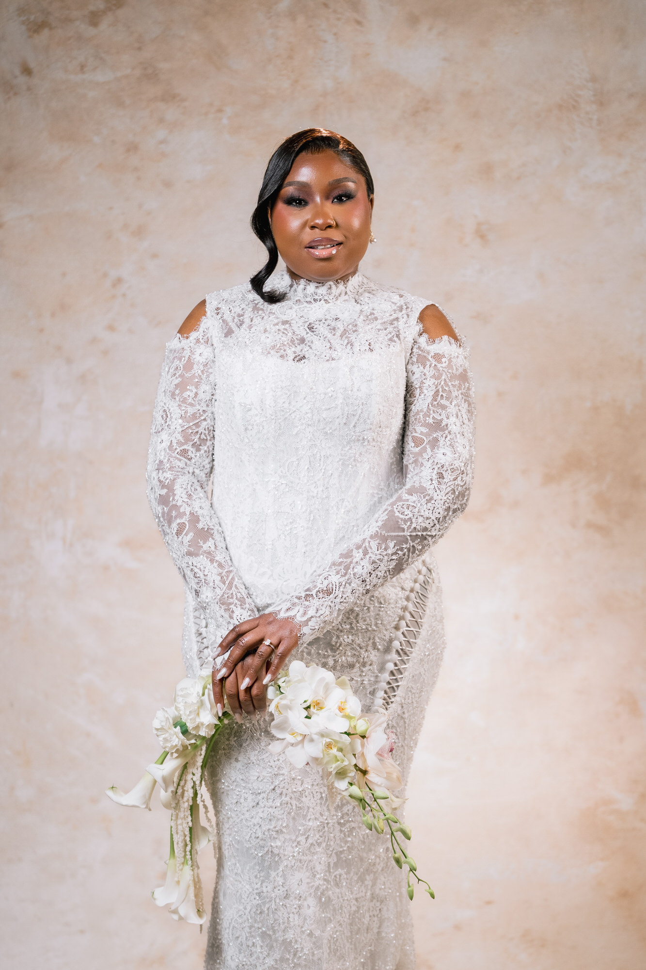 Bride in a white lace wedding dress with cutout shoulders, holding a white bouquet, posed against a beige textured backdrop.