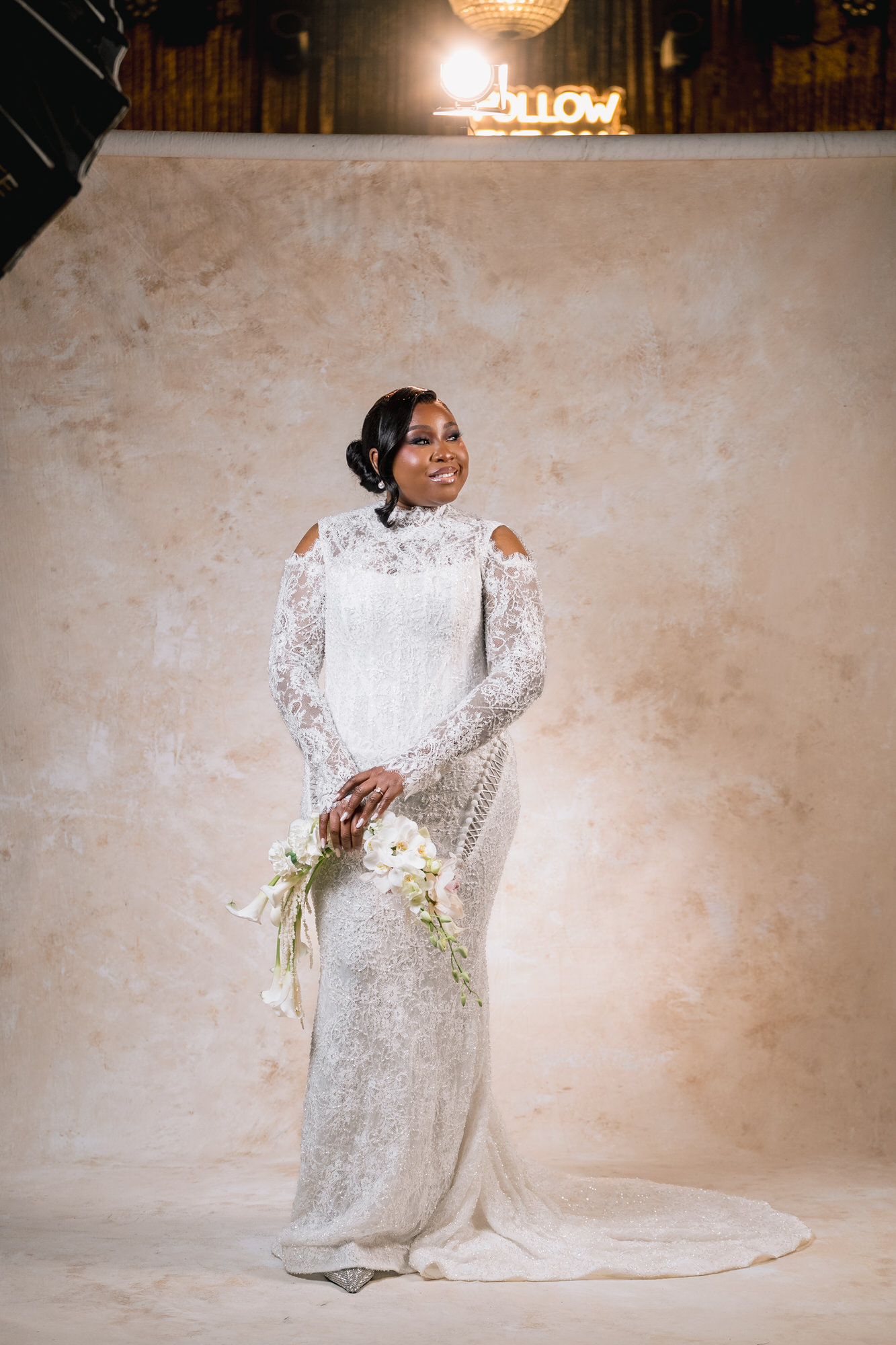 Bride in a white lace wedding gown holding a cascading bouquet, standing against a beige studio backdrop.