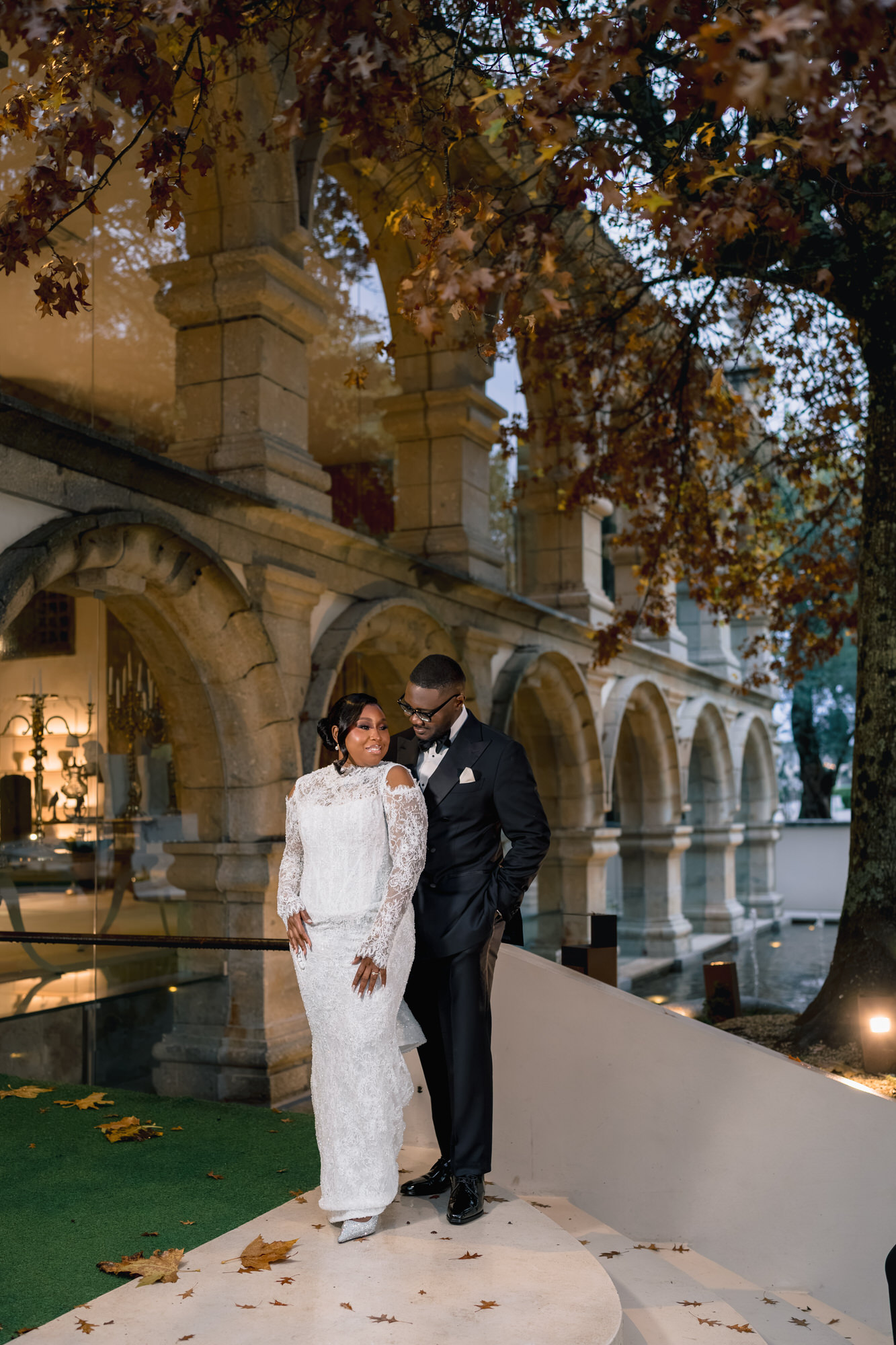Bride in a white lace gown stands with a groom in a black suit on an arched stone staircase under autumn leaves.