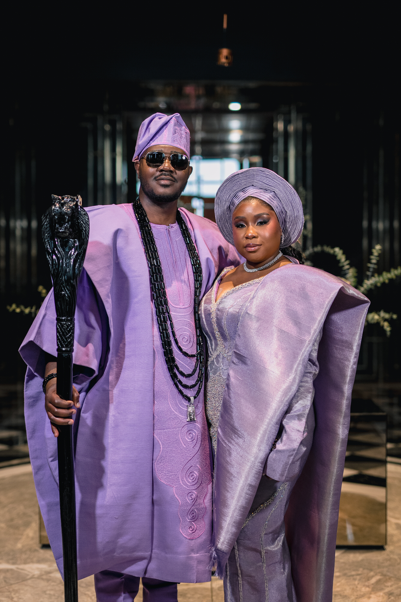 A man and woman in matching lavender traditional outfits, the man holding a staff and wearing sunglasses indoors.