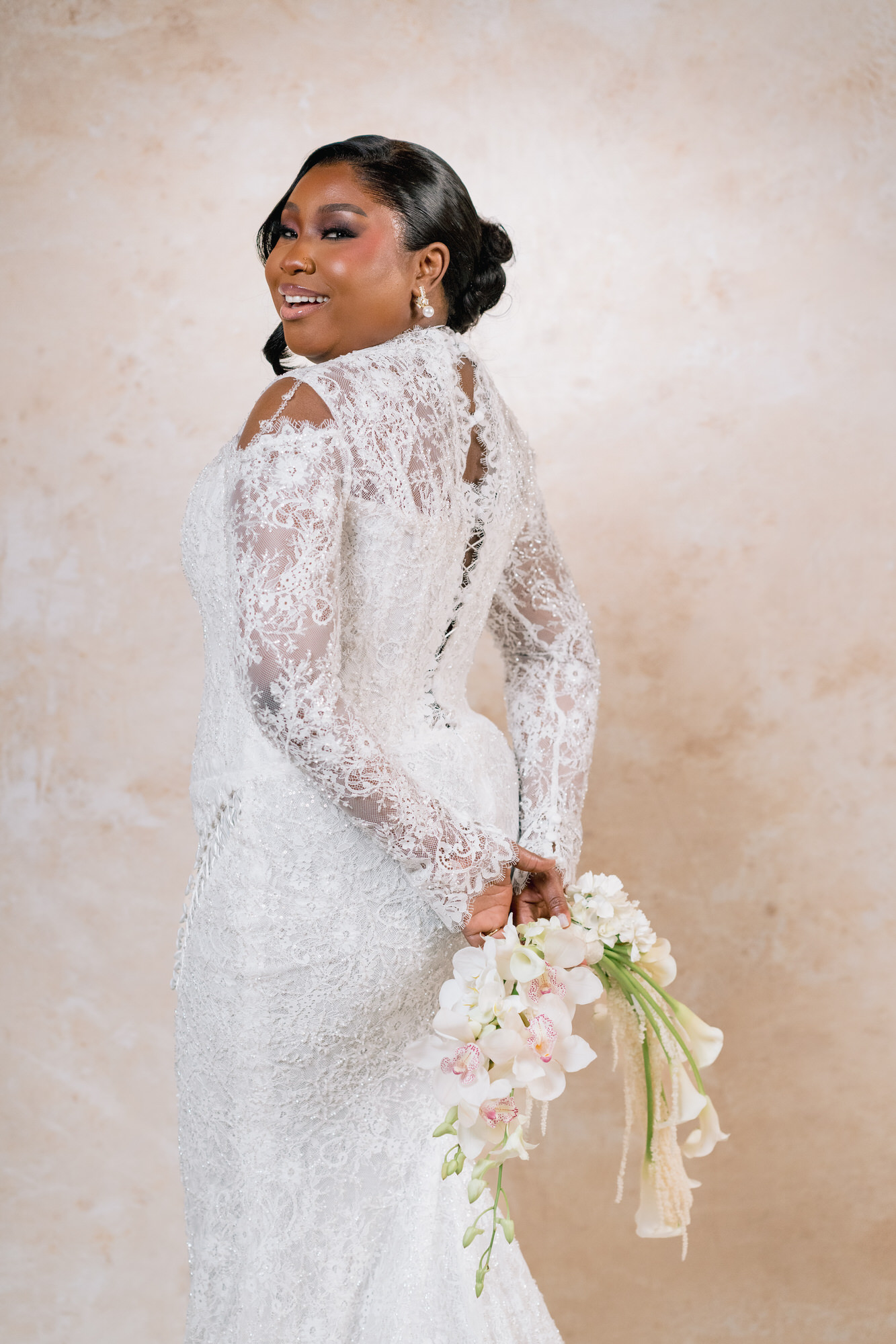 Bride in a white lace wedding gown, turned slightly to the side, smiling and holding a cascading bouquet of white flowers.