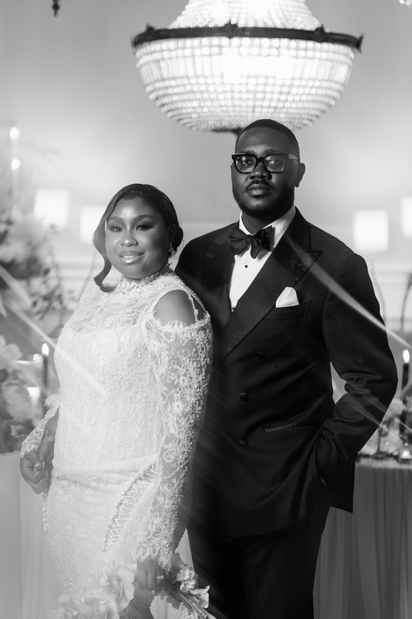 Bride in a lace wedding gown and groom in a black tuxedo posing indoors beneath a grand chandelier, black-and-white photo.