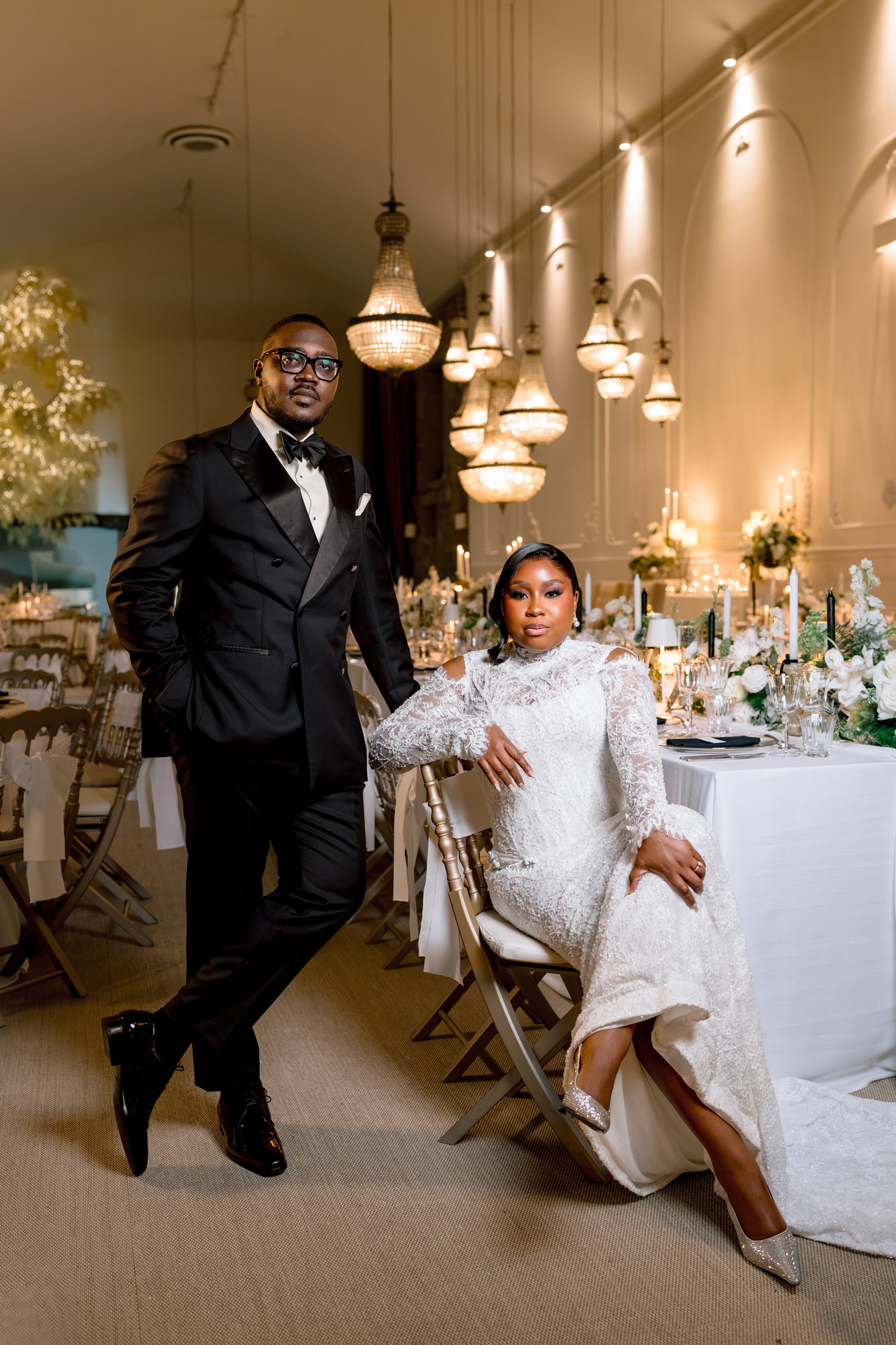 Man in a black tuxedo stands beside a seated woman in a white lace gown at a chandelier-lit wedding reception hall.
