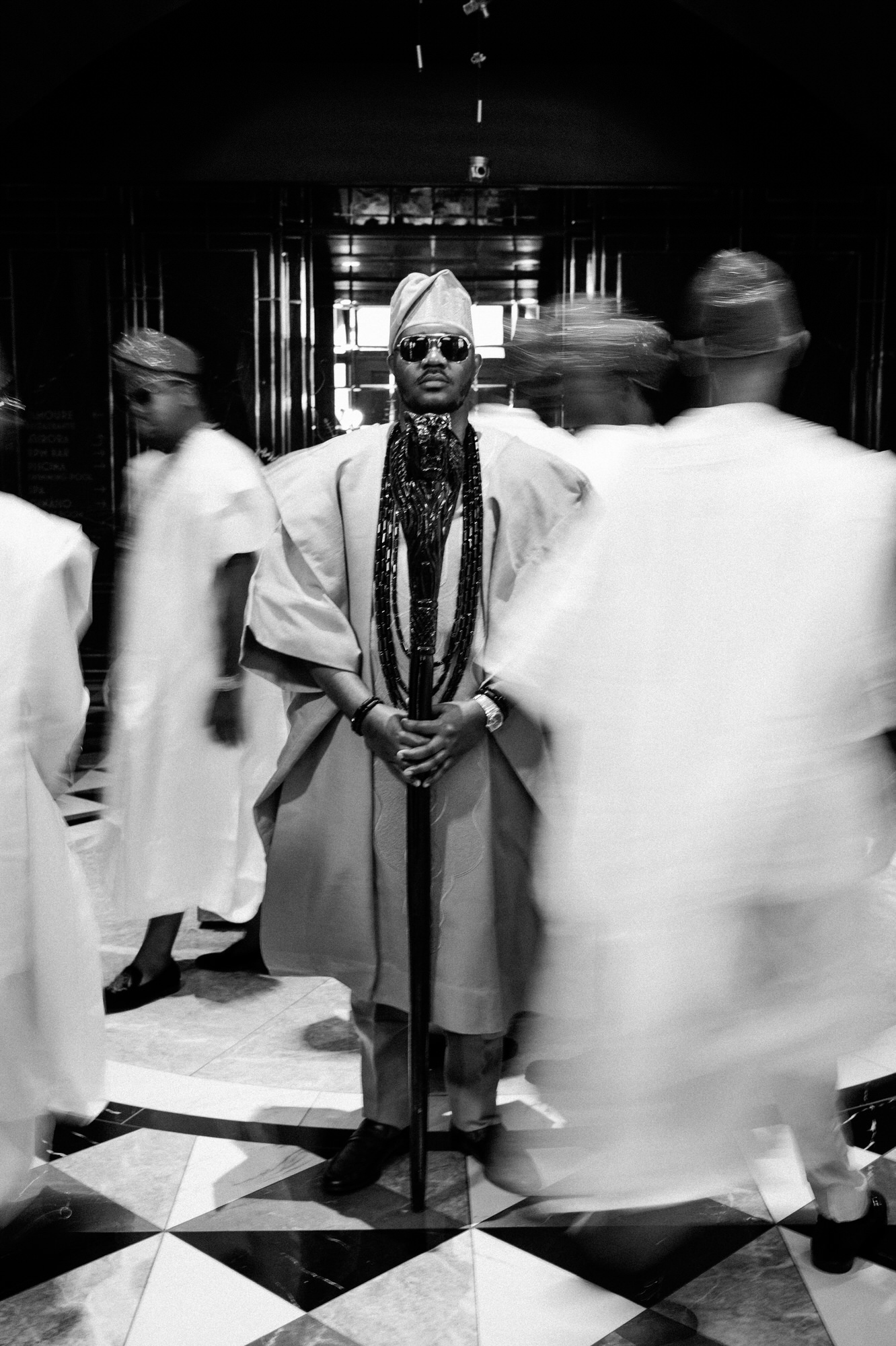 Man in traditional robes and hat, wearing sunglasses and multiple bead necklaces, standing with a staff as blurred figures move around him in a tiled, reflective space.