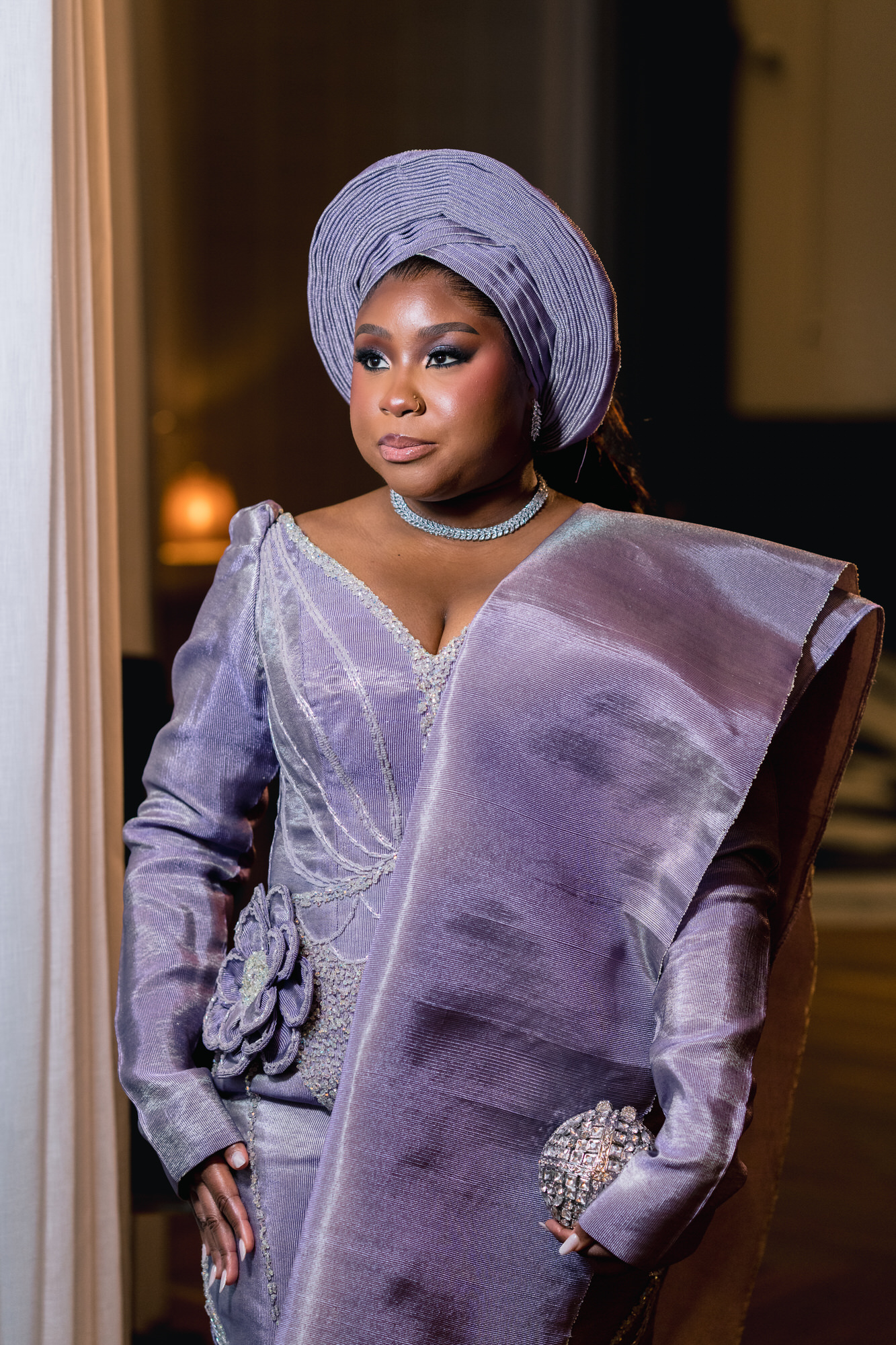 Woman in lavender traditional outfit with a matching headwrap, standing beside a curtain in a softly lit room.