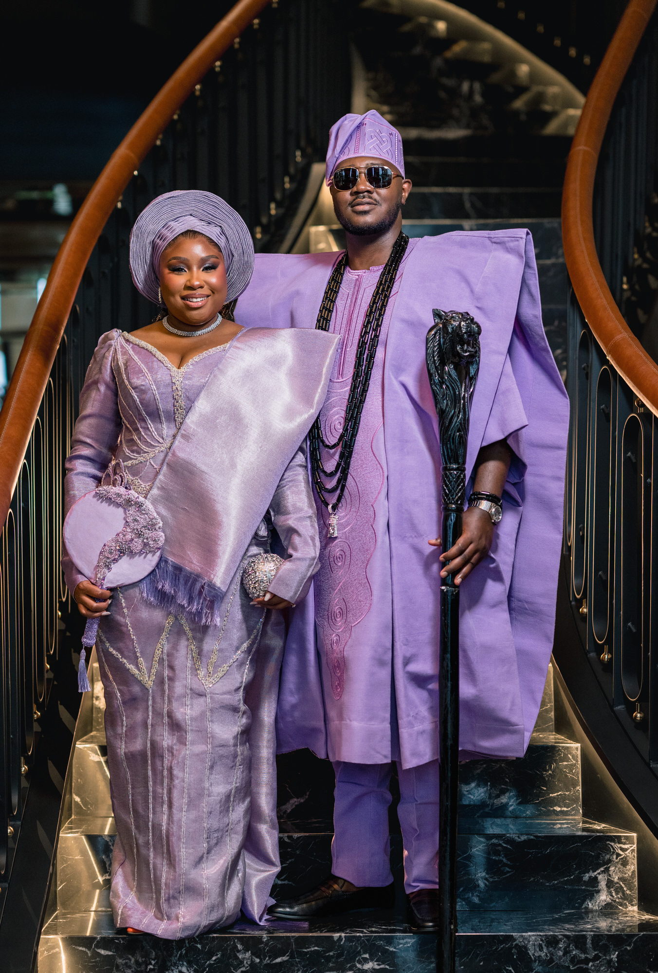 Smiling couple in lavender traditional outfits standing on a marble staircase, woman with a large lavender head wrap and flowing gown, man in a matching purple agbada holding a staff.