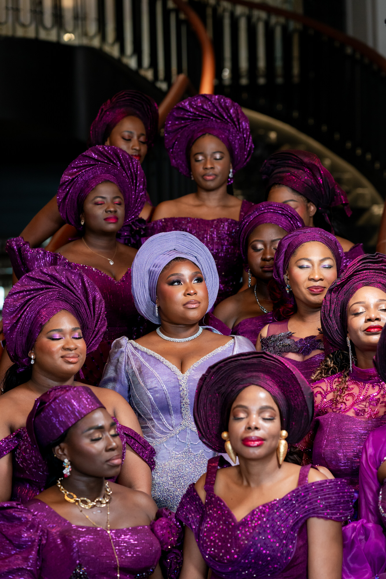 Group of women in glittery purple outfits and headwraps posing on a staircase, center woman in lavender dress stands out with a silver headwrap.