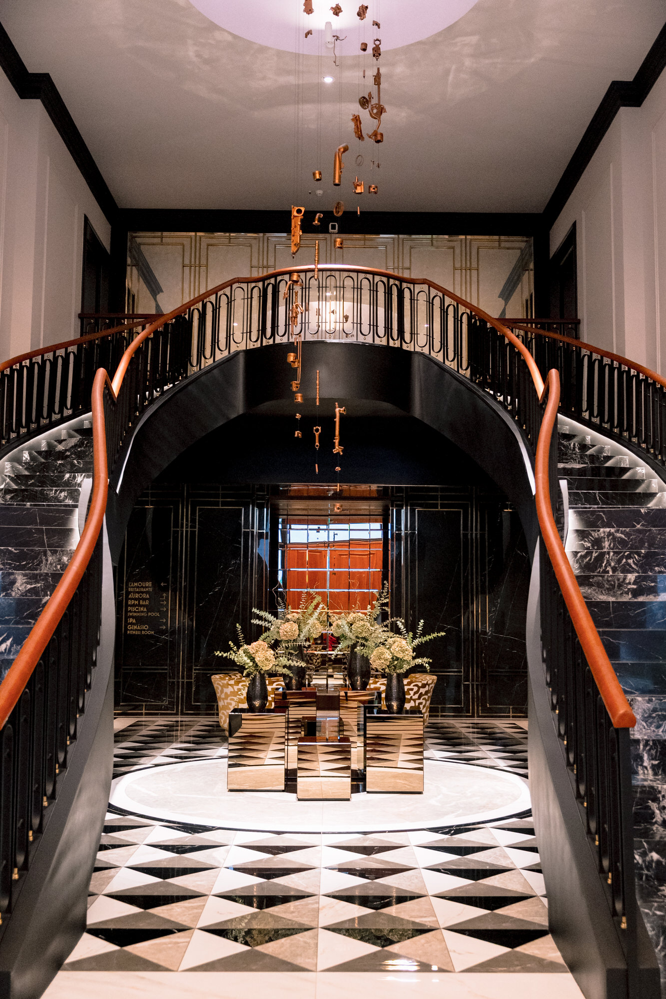 Grand lobby staircase with black railing and copper hanging sculpture above, set on a black-and-white checkered marble floor with floral arrangement nearby.