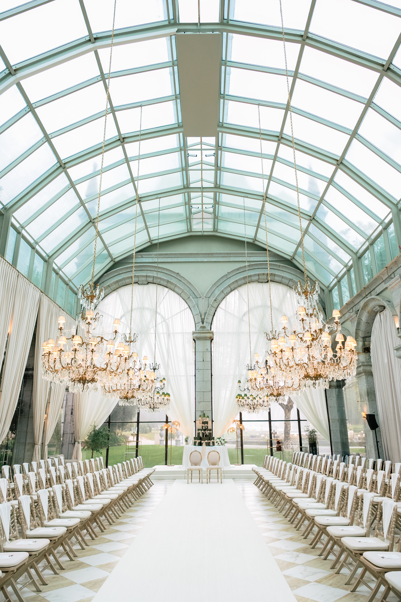 Elegant indoor wedding venue with a glass-domed ceiling, chandeliers, and rows of white chairs along a central aisle.