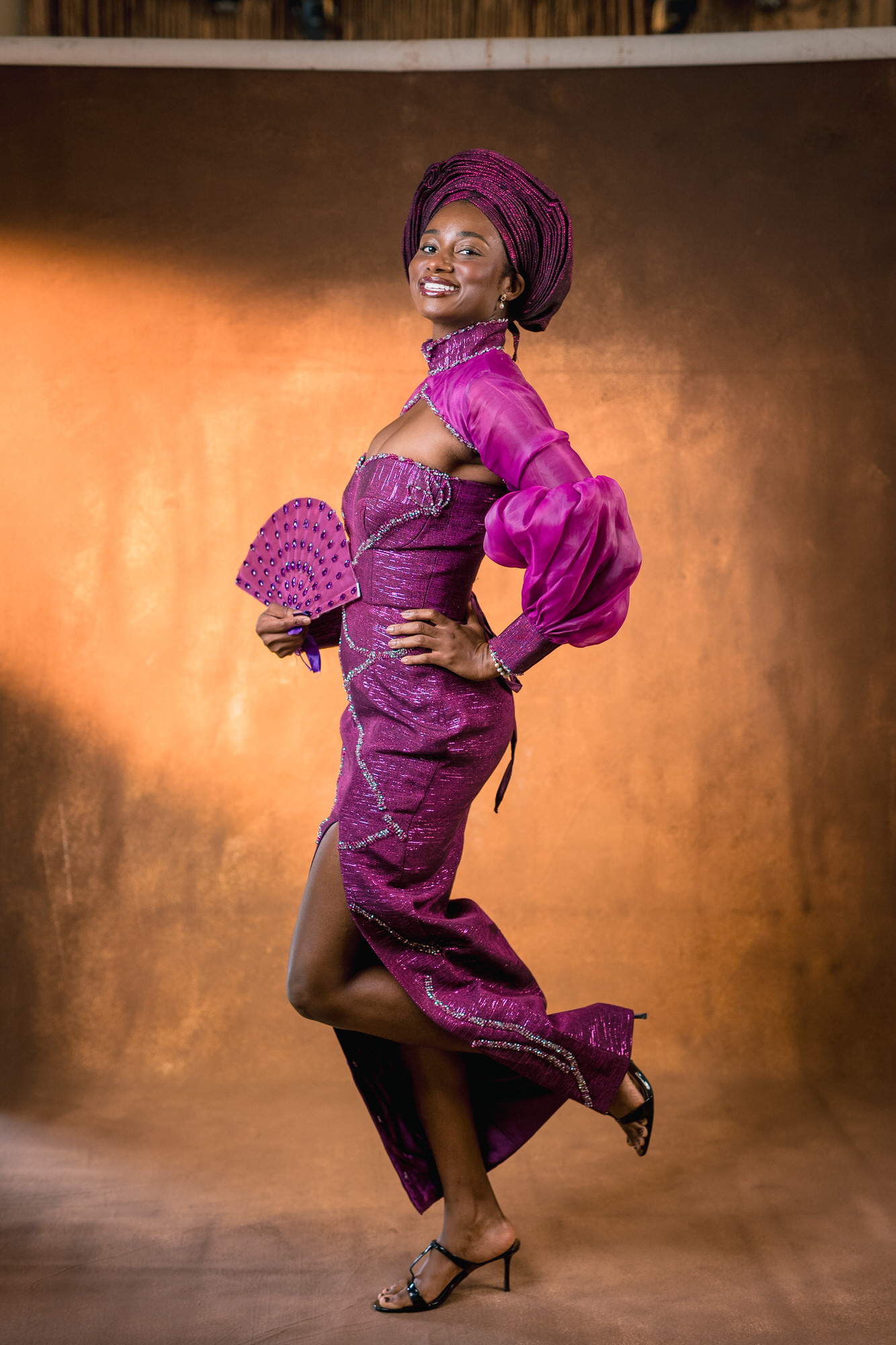 Woman in a sparkling magenta dress and matching headwrap poses mid-dance, holding a decorative pink fan against a bronze backdrop.