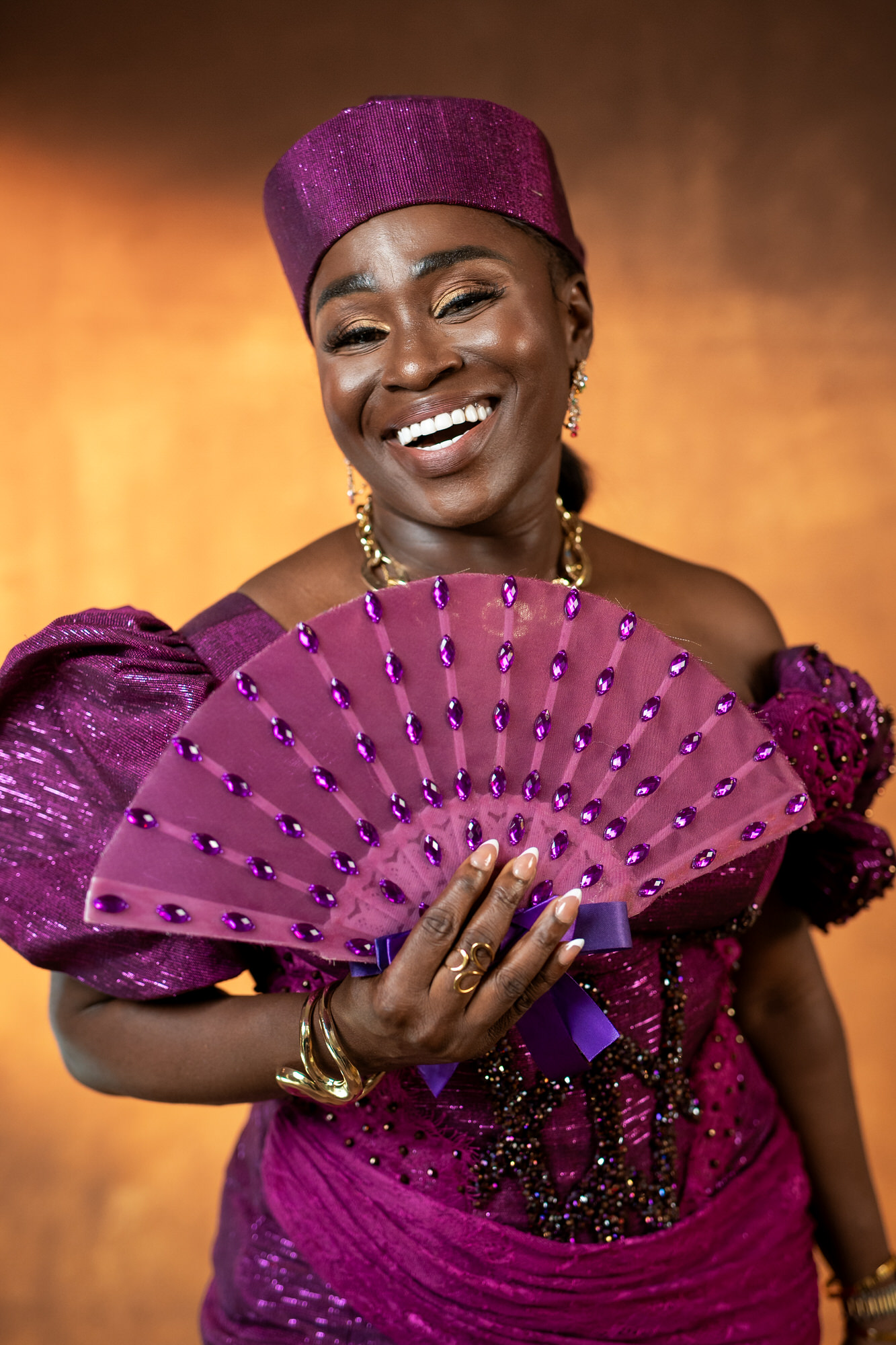 Smiling woman in a glittering purple gown and headwrap, holding a pink jeweled fan against a warm amber background.