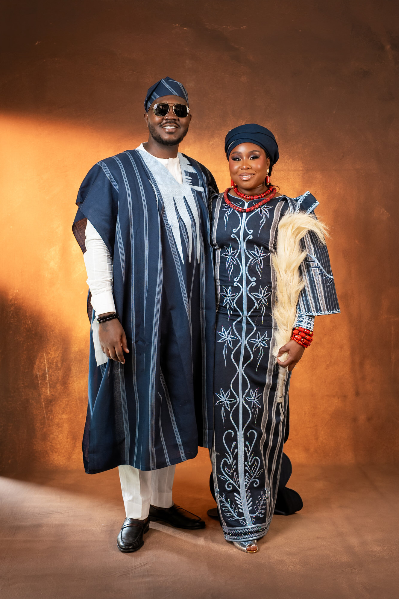 A couple in matching blue traditional outfits posing together against a warm brown backdrop, smiling at the camera.