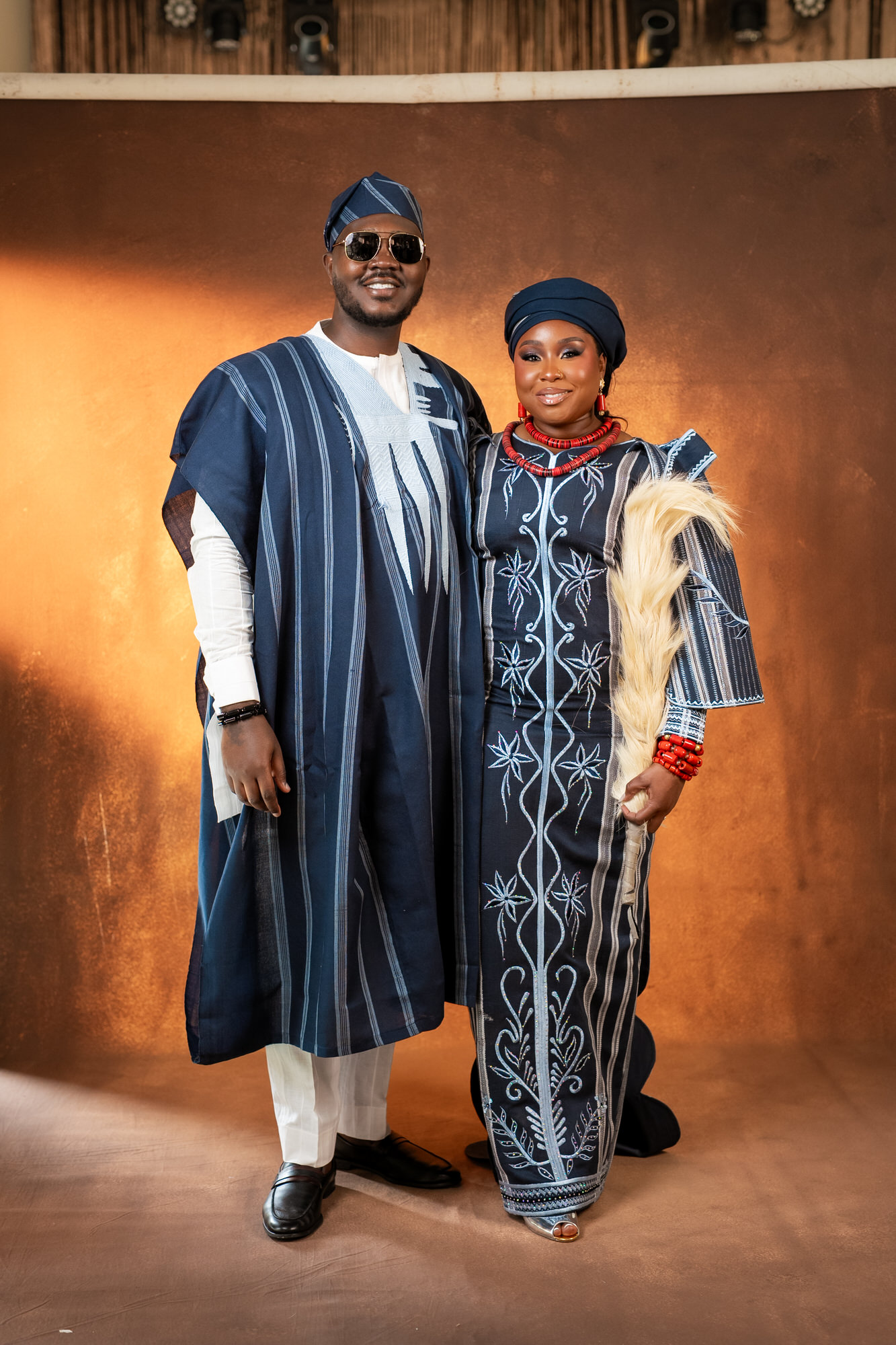 Couple in matching blue traditional outfits smile for the camera indoors against a warm backdrop
