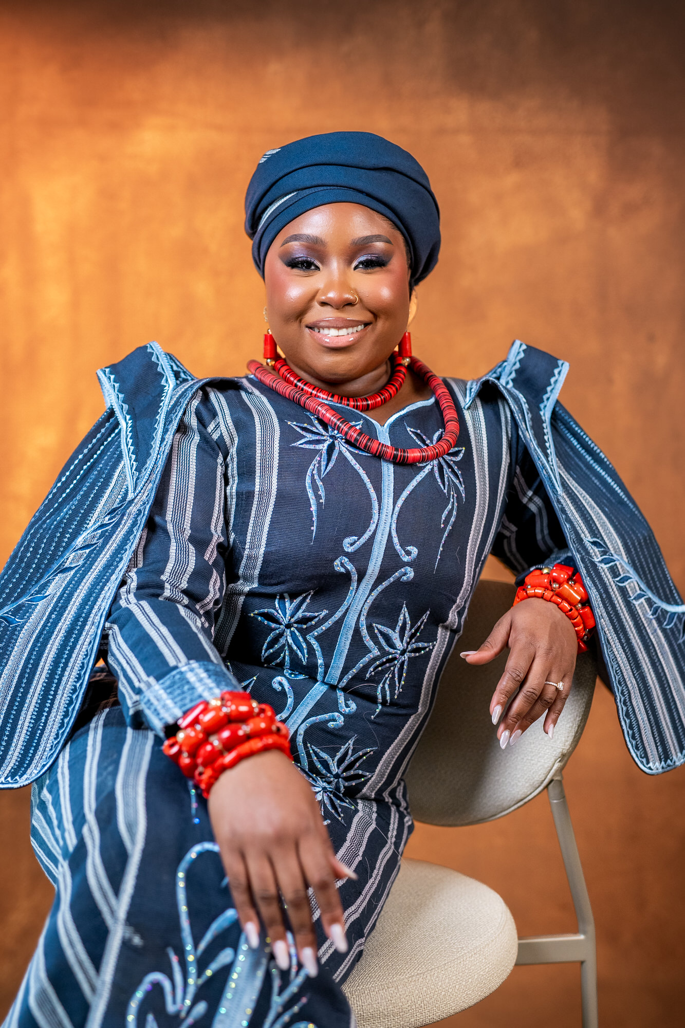 Smiling woman in a blue patterned traditional outfit with a head wrap and red beaded necklace, seated against a warm brown backdrop.