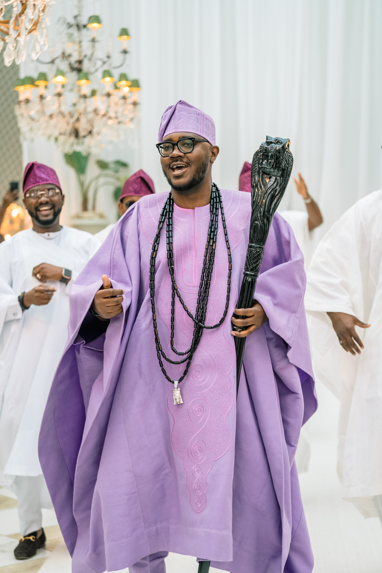 Five adults posing for a celebration photo indoors, wearing matching purple traditional outfits with head wraps and beads, beneath a crystal chandelier and green-and-white checkered floor.