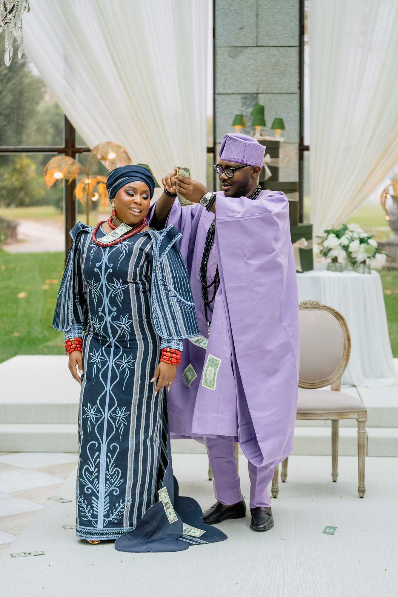 Couple in traditional attire celebrating, man in lavender robe hands woman money while dollar bills float around them at a festive event.