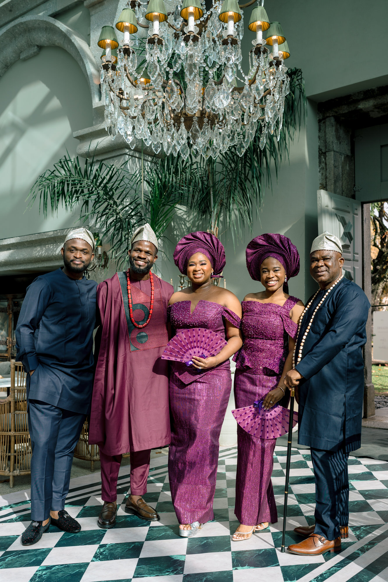 Five adults posing for a celebration photo indoors, wearing matching purple traditional outfits with head wraps and beads, beneath a crystal chandelier and green-and-white checkered floor.