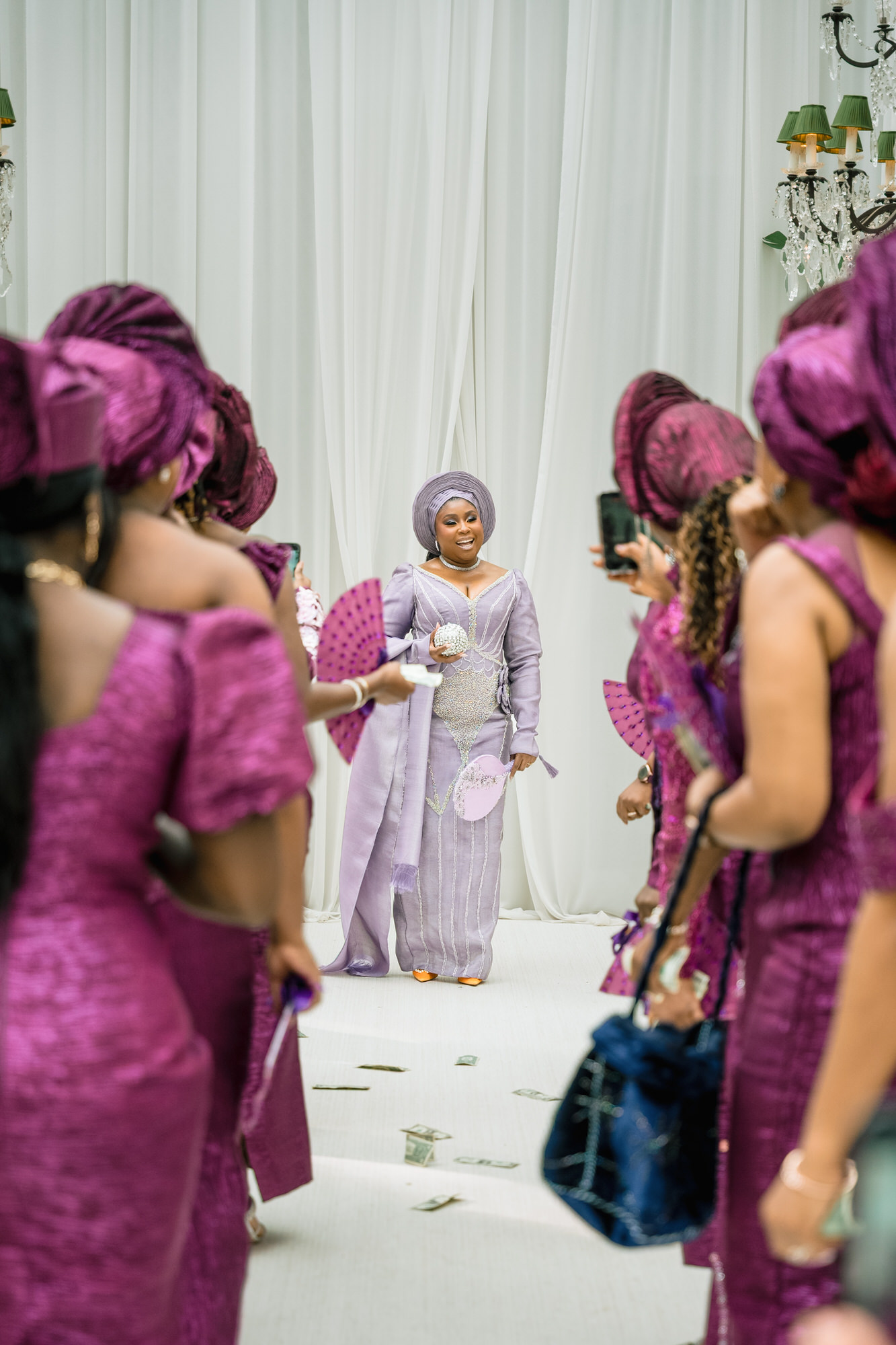 Woman in a lavender traditional dress and headwrap walking down a white aisle, surrounded by guests in matching purple outfits; dollar bills scattered on the floor.