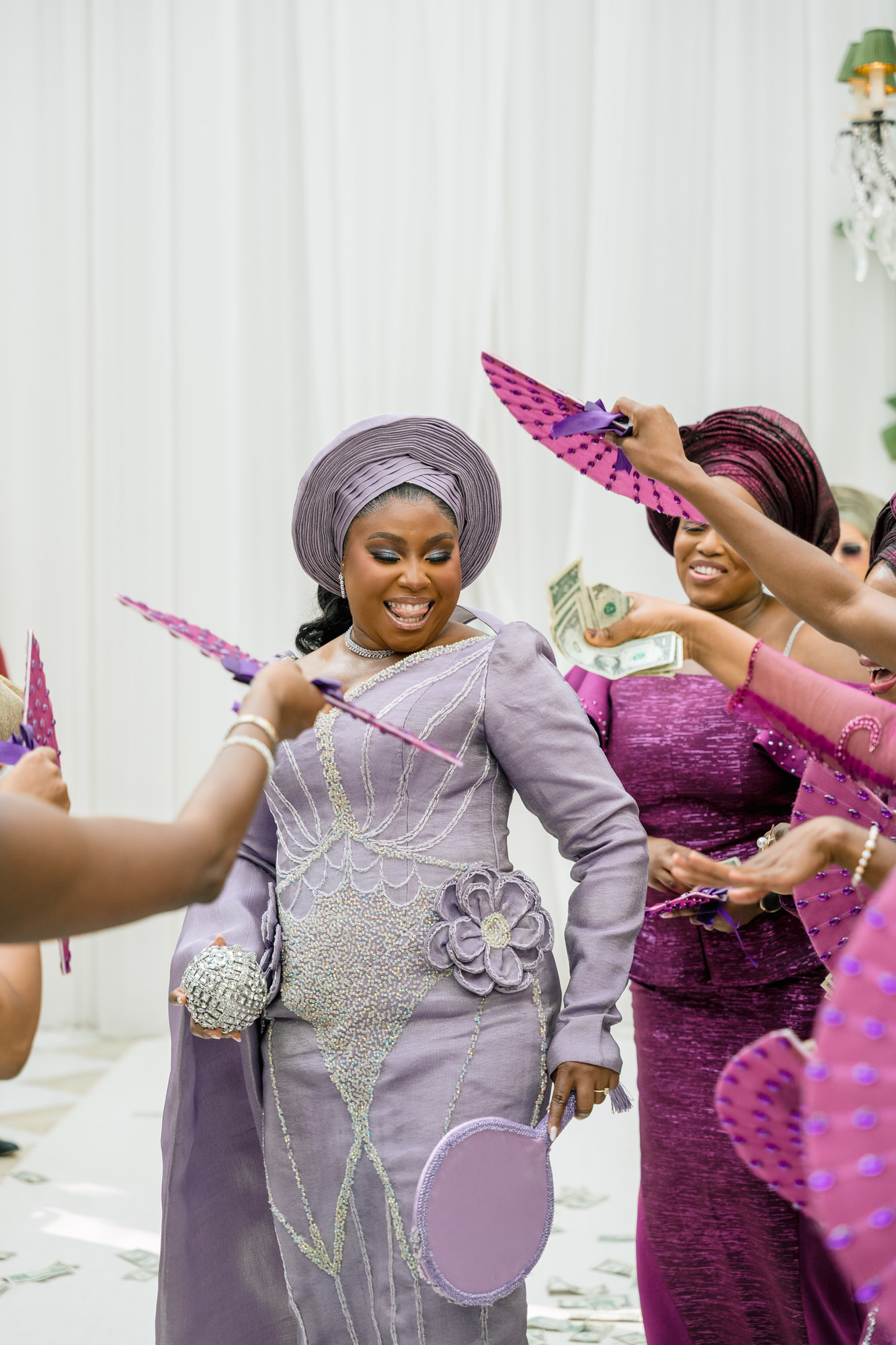 Joyful woman in a lavender traditional dress and headwrap dances while friends in purple throw money during a celebration, green bills in the air/hand.