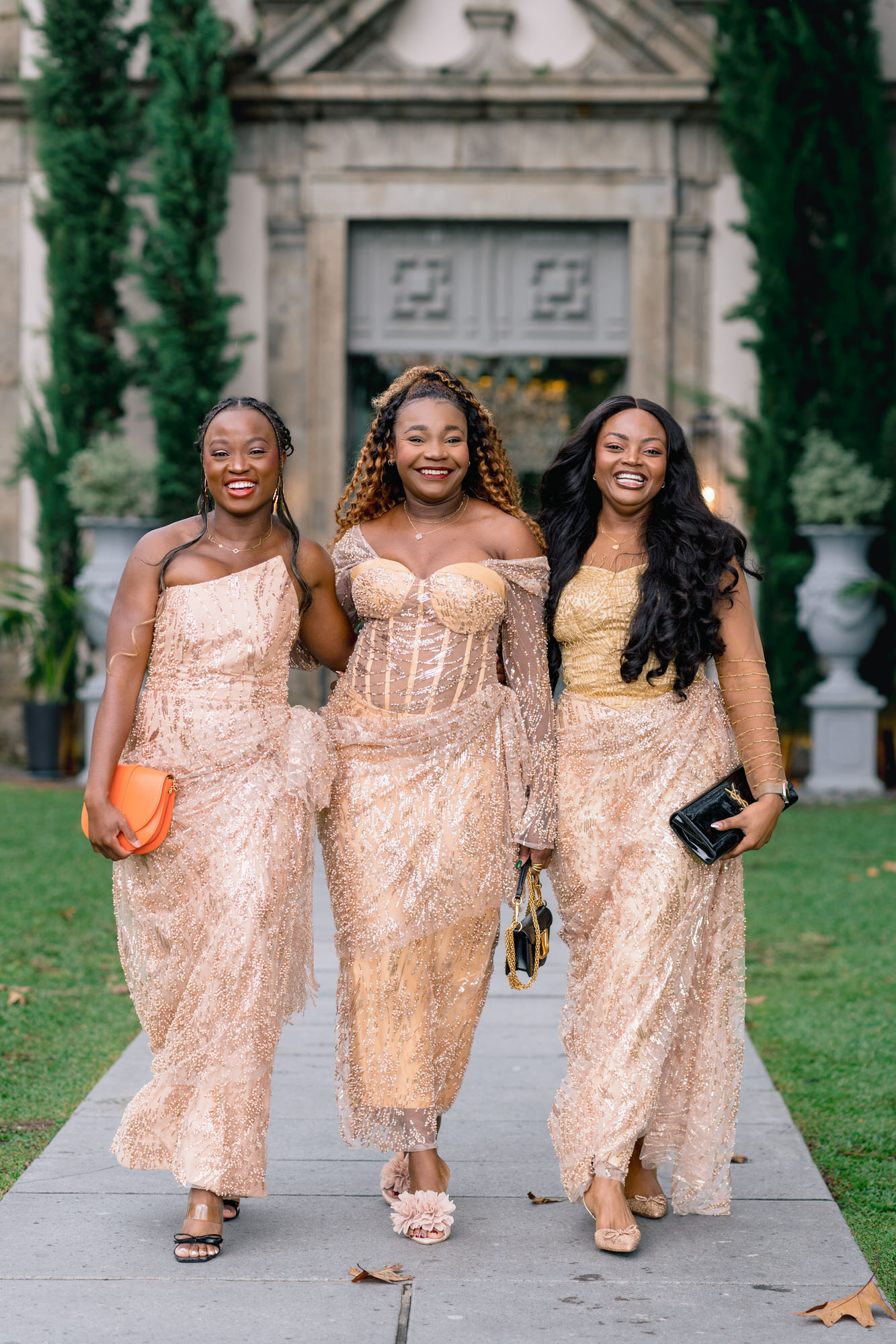 Three women in matching champagne sequin dresses walk together, smiling, on a stone path outside a building with greenery in the background.