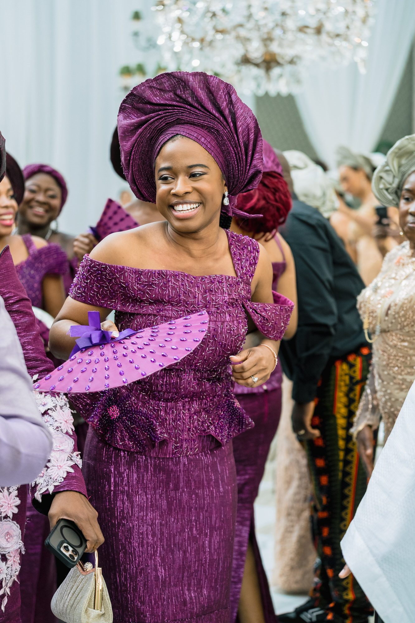 Smiling woman in a purple traditional outfit and head wrap at a festive gathering.