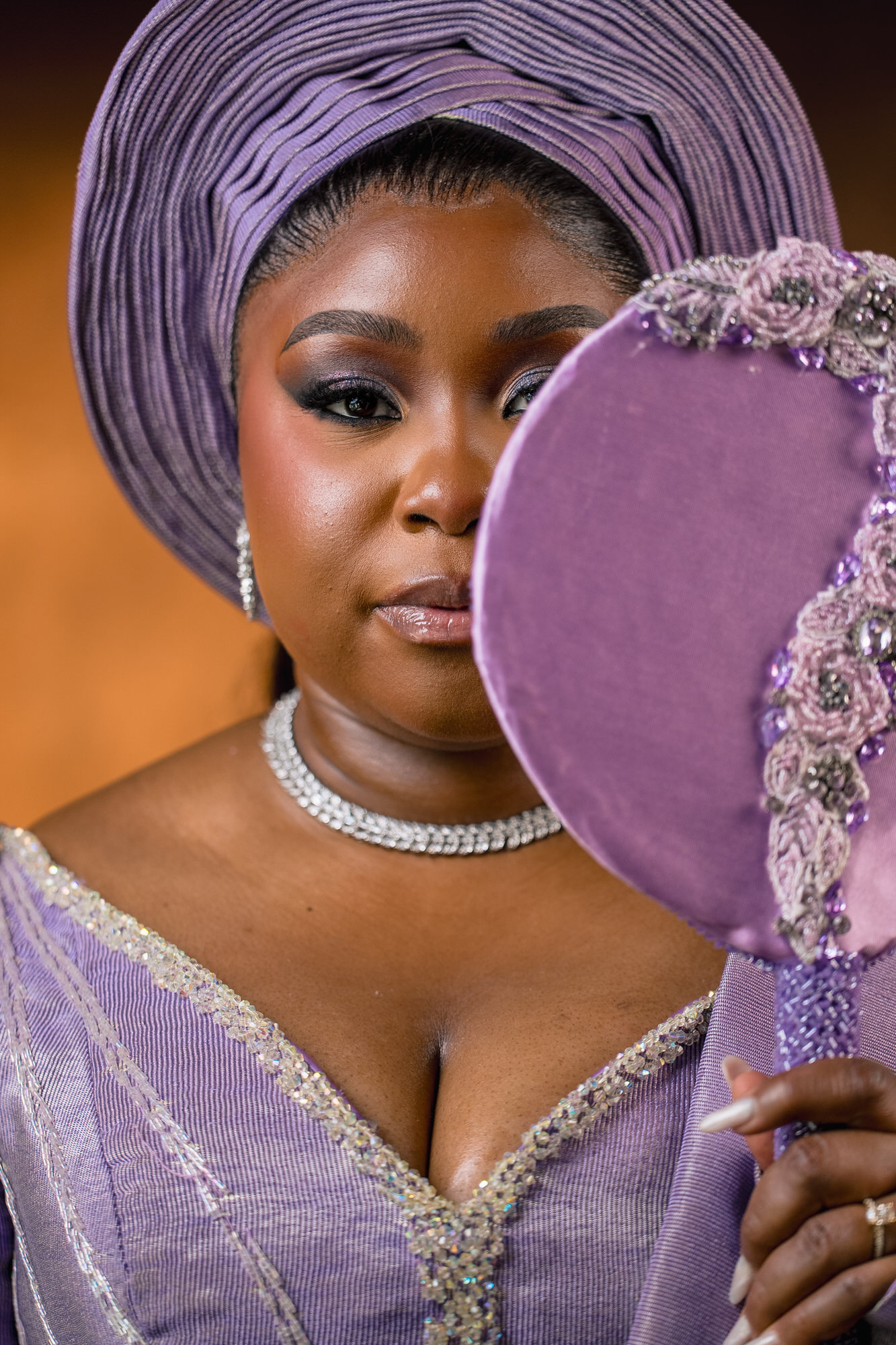 Close-up of a woman wearing a purple gele and matching embellished dress, showing smooth makeup and silver jewelry.