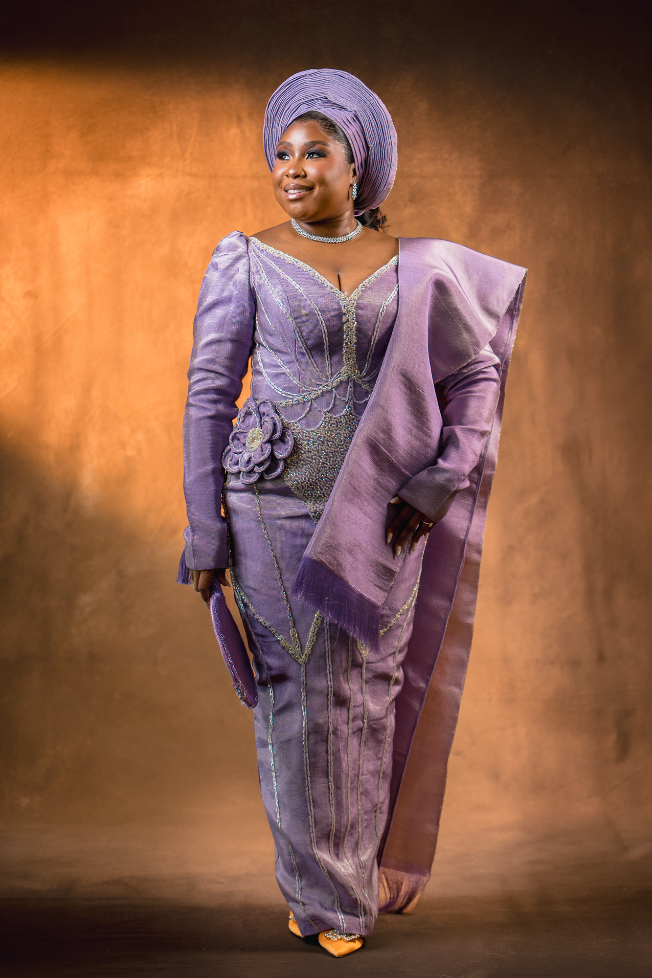 Woman in a lilac traditional gown with a matching head wrap, standing on a stage with a warm backdrop.
