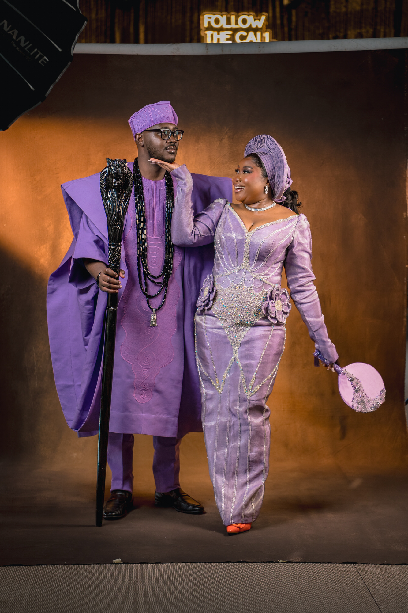 Couple dressed in matching purple traditional outfits posing on a stage; man holds a ceremonial staff and wears a hat, woman smiles in a beaded purple gown with headwrap and holds a decorative prop umbrella-shaped accessory in the background is a warm, backlit set.