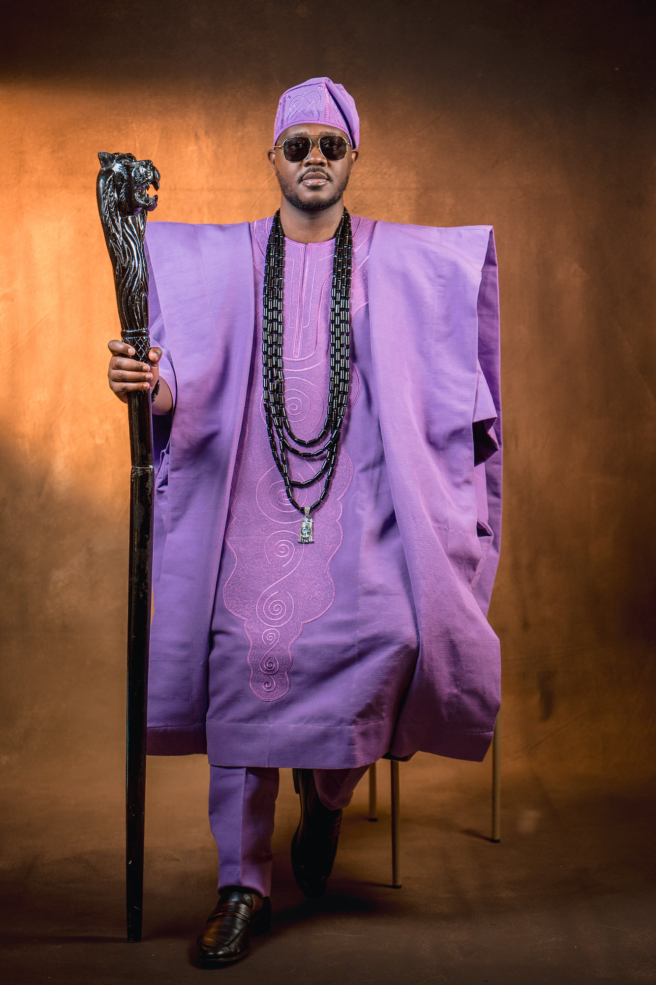 Man in a purple traditional outfit and hat, holding a tall staff with a carved top, wearing sunglasses against a warm background.