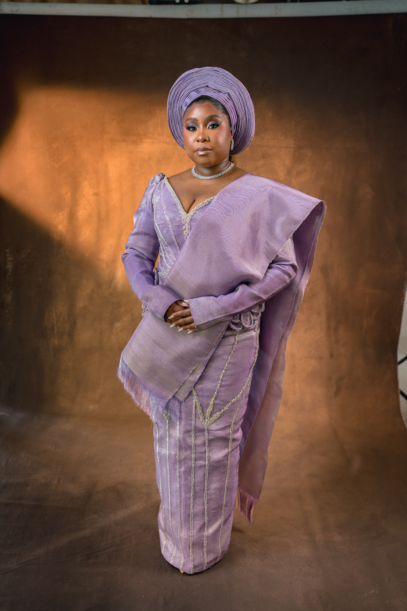 Woman in a lavender traditional outfit and head wrap standing with hands clasped against a bronze studio backdrop.