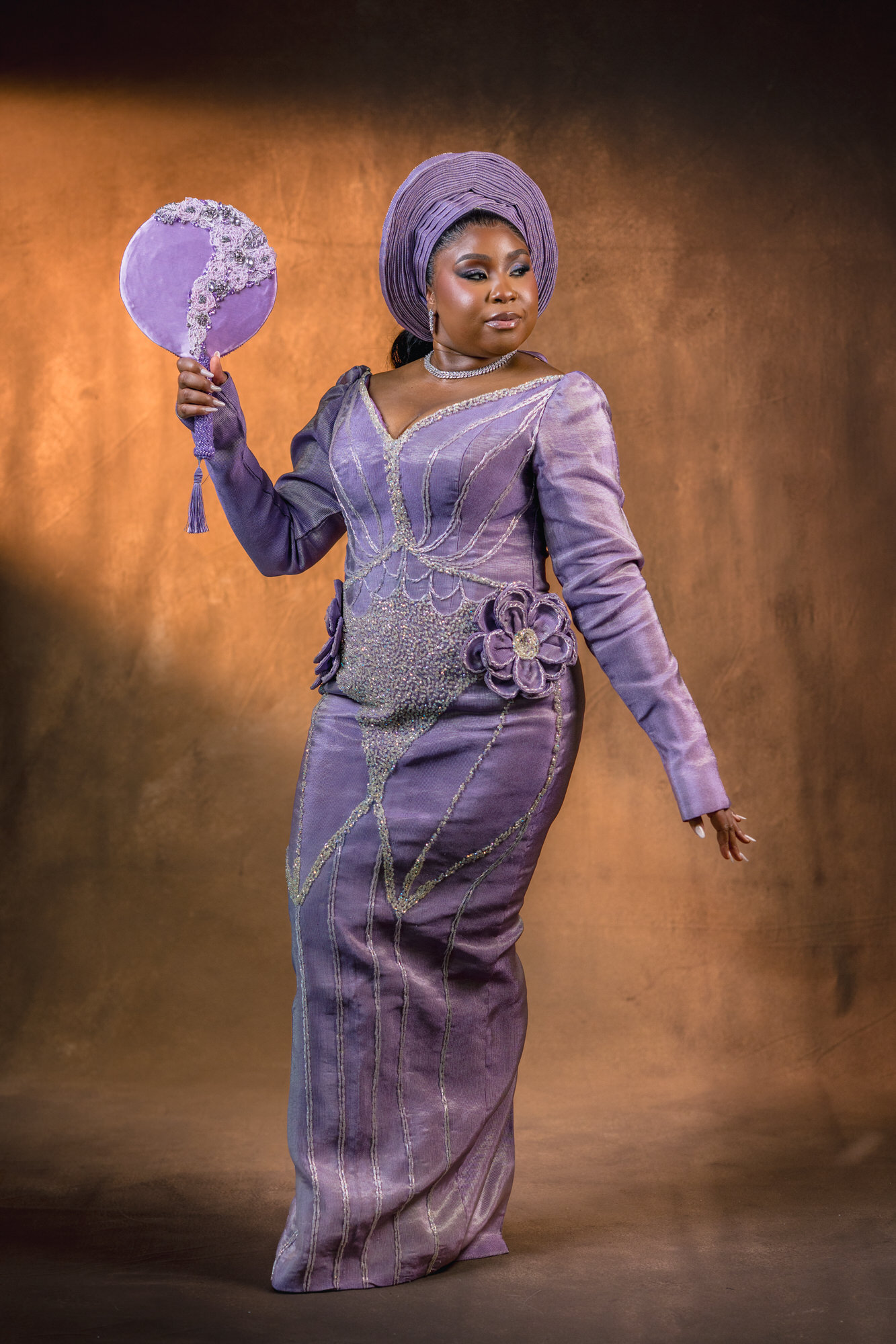 Woman in a lavender gown and matching headwrap, holding a circular purple fan, against a warm brown backdrop.