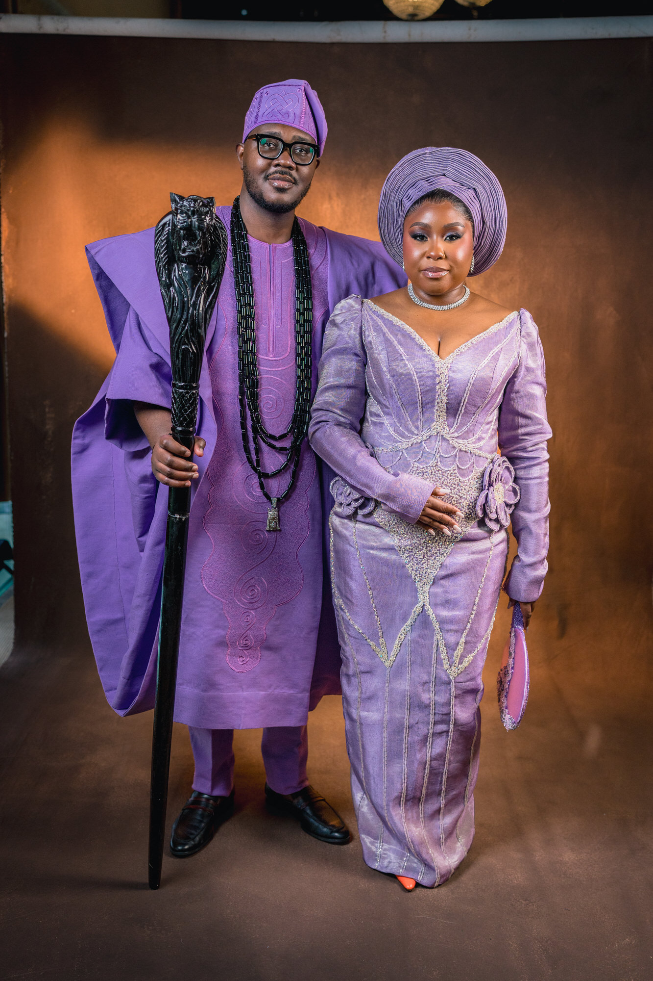 Couple in matching lavender traditional outfits: man with a carved staff and beaded necklaces, woman in a lavender dress with a purple head wrap and jewel necklace, posing together.