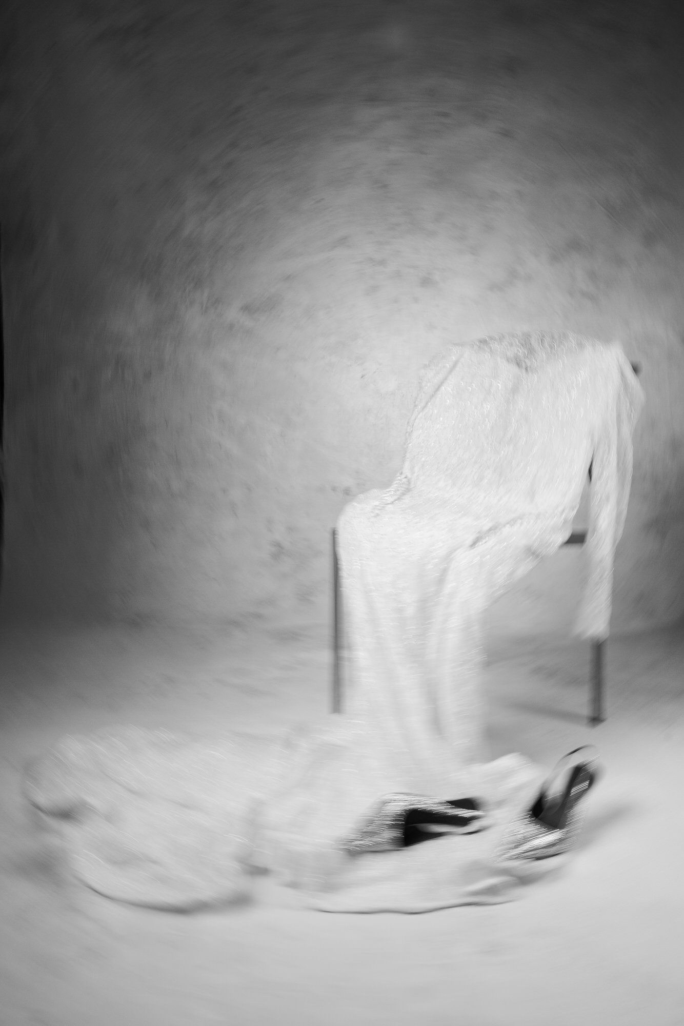 Black-and-white image of a person wearing a shimmering, back‑facing gown, seated near a chair with a soft blur around.